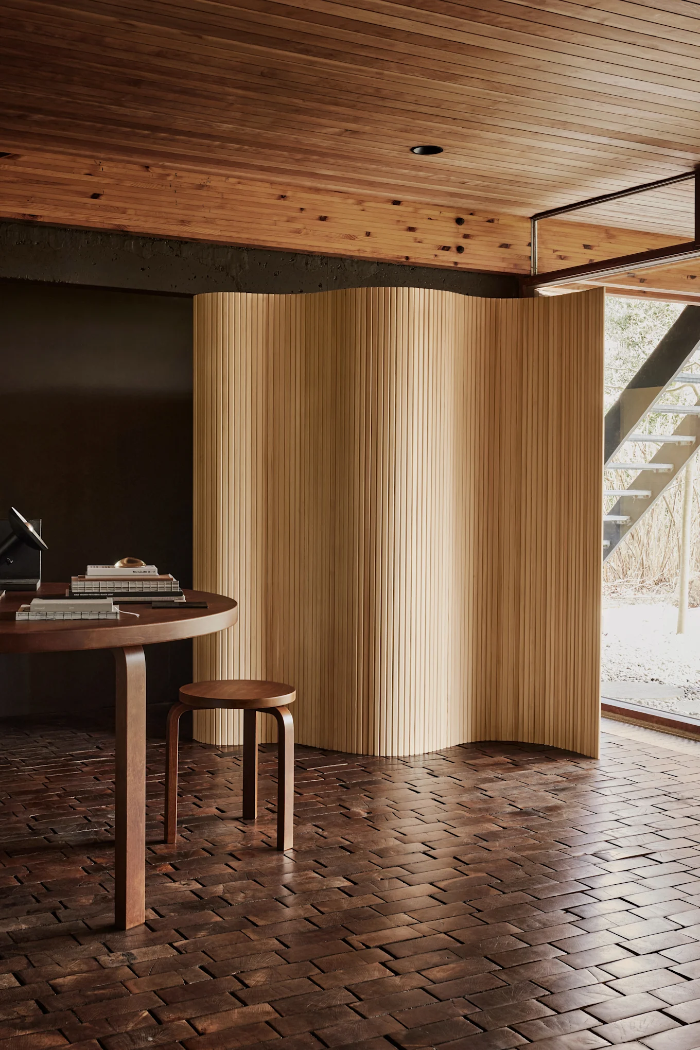 Modernes Zimmer mit einem runden Holztisch, einem kleinen Hocker und einem wellenförmigen Holzschirm. Dunkler Holzblockboden, helle Holzlatten an der Decke und ein Fenster zu einer Außentreppe.