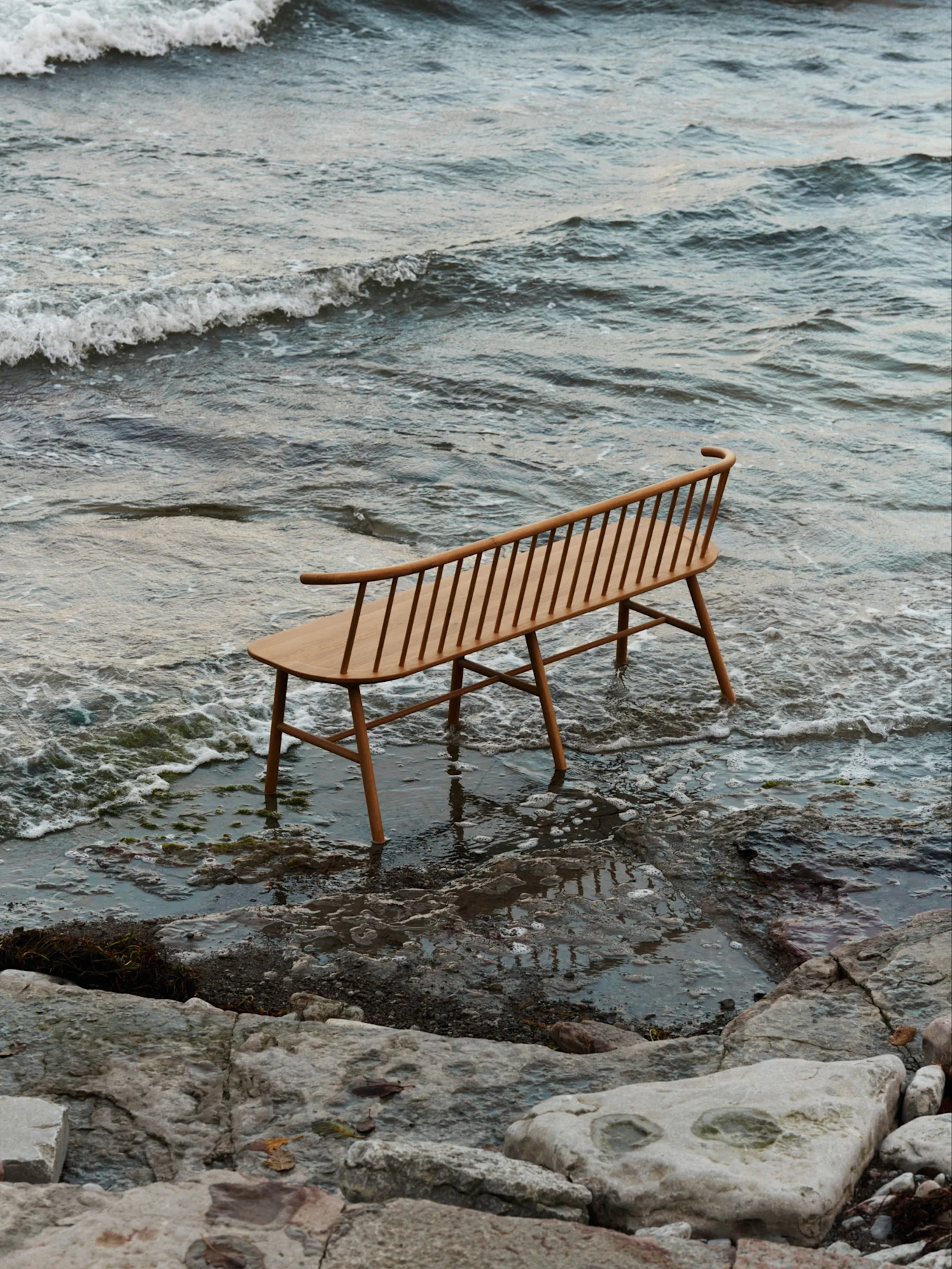 Eine helle Holzbank mit Latten und geschwungener Rückenlehne steht im seichten Wasser an einem felsigen Strand mit sanften Wellen.