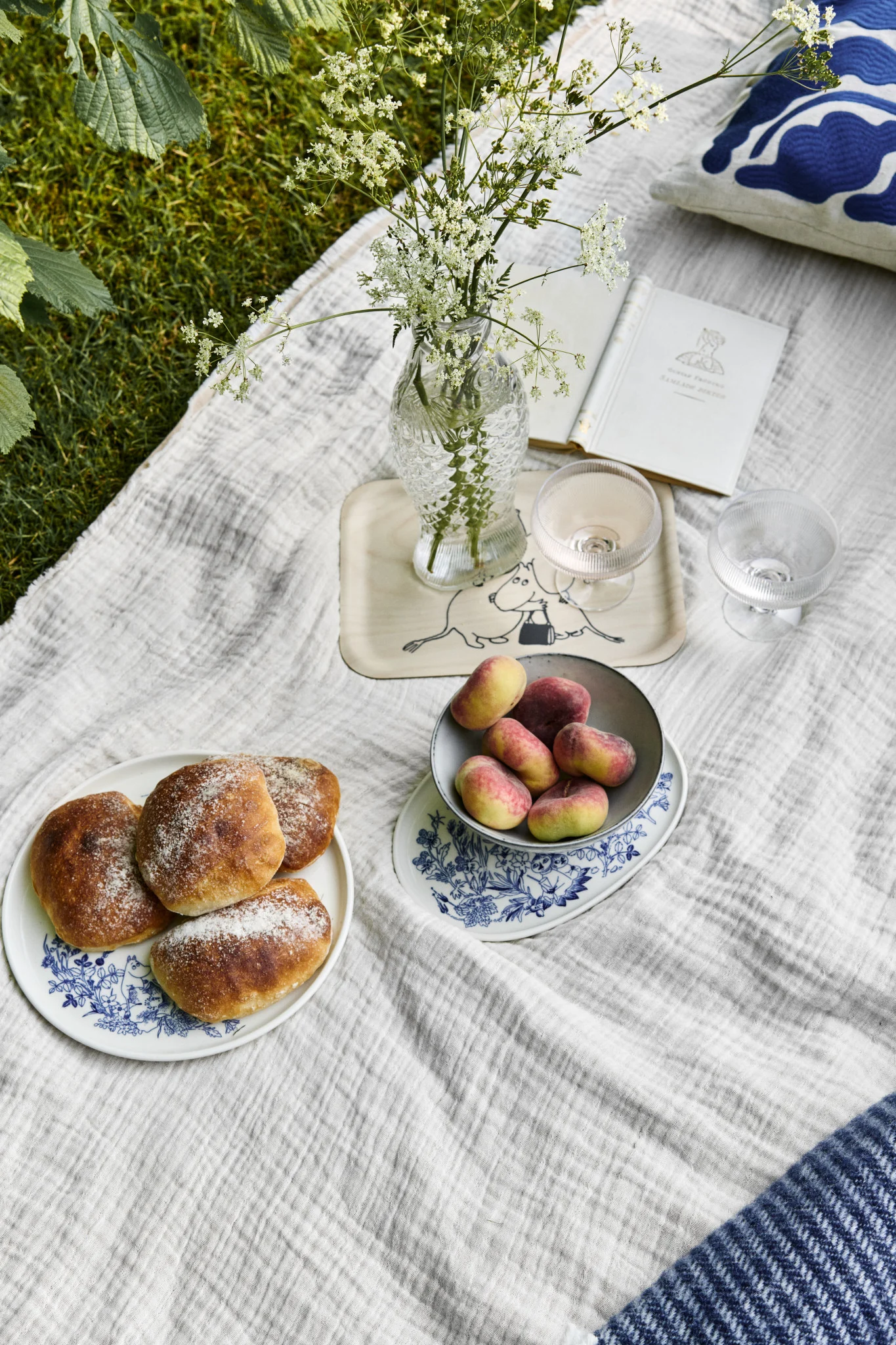 Picknick auf Gras mit Brötchen, Plattpfirsichen, Wildblumen in Vase und offenem Buch auf weißer Decke.