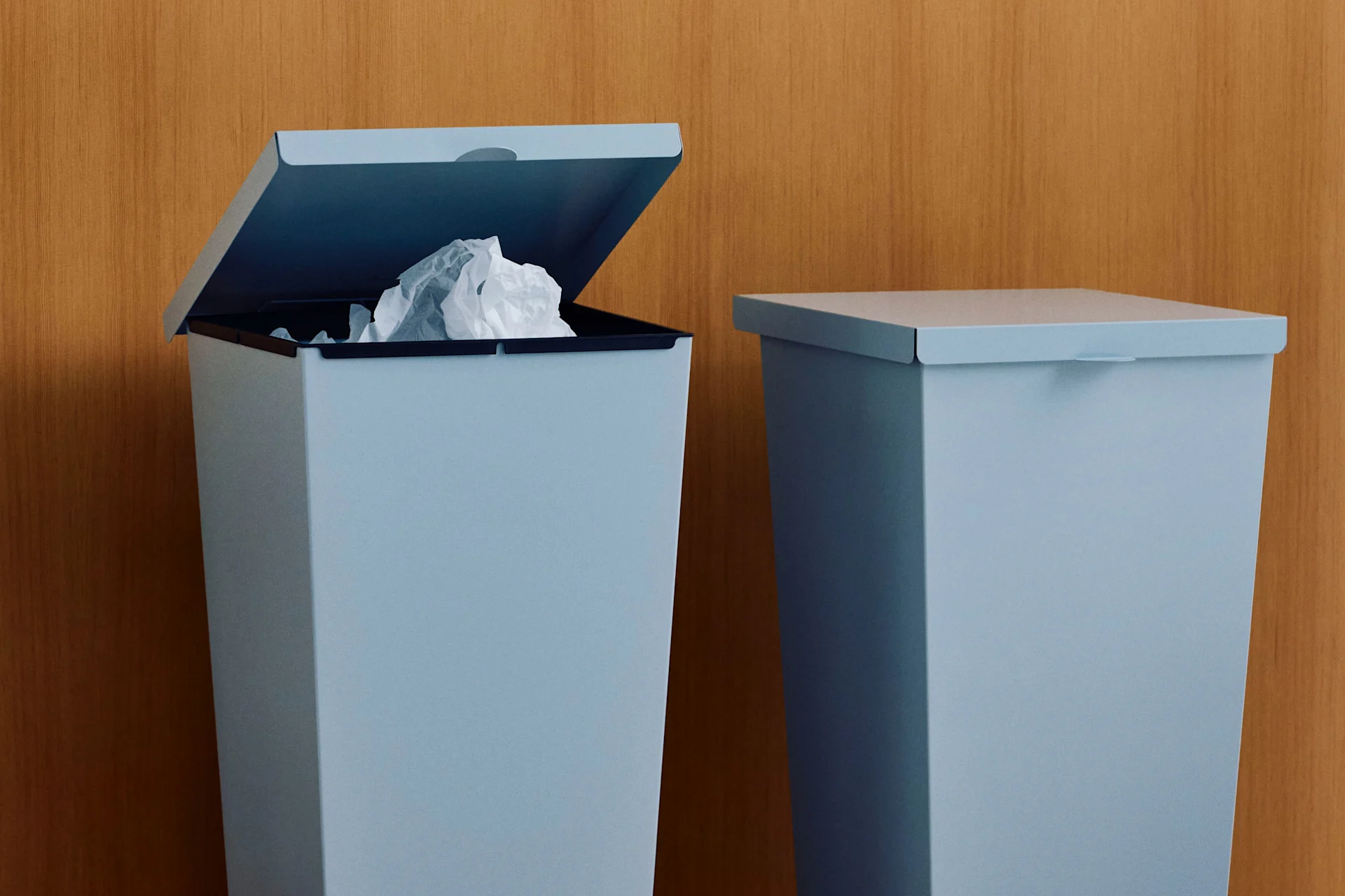 Zwei hellblaue Mülleimer vor einer Holzwand, einer offen mit zerknülltem Papier.