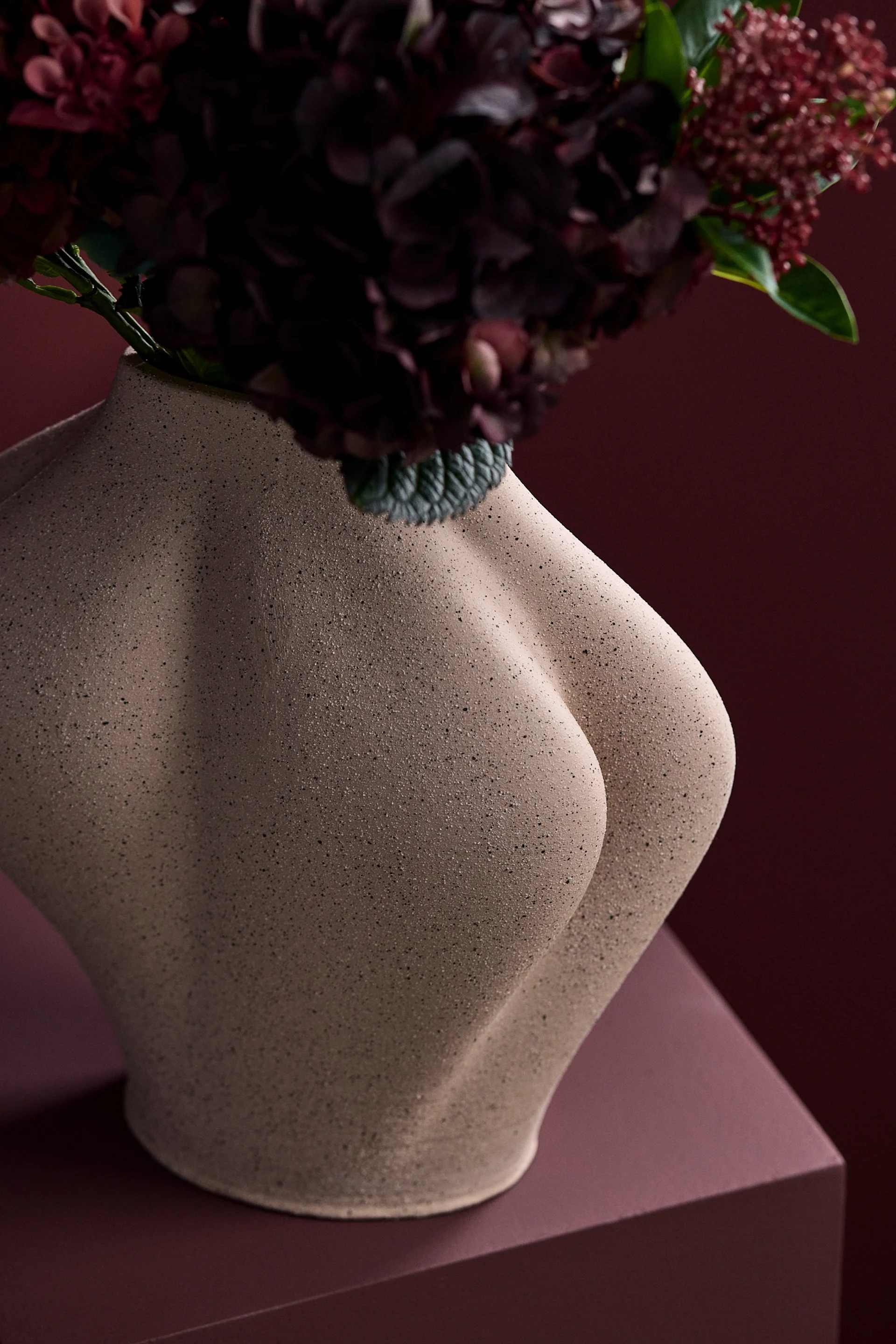 Skulpturale beige Vase in Form eines menschlichen Torsos, mit dunkelroten Blumen vor einem passenden roten Hintergrund.
