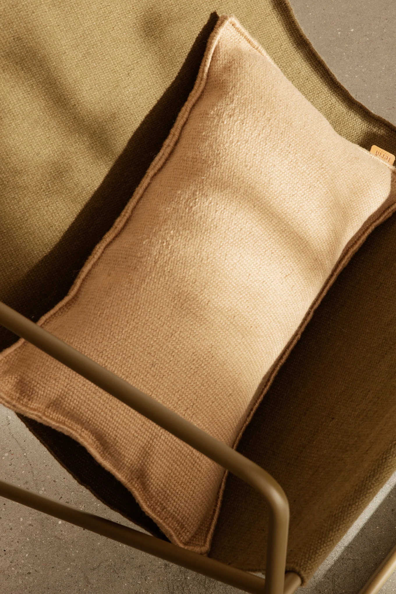 Draufsicht einer hellbraunen Auflage auf einem dunkelolivgrünen Stuhl aus strukturiertem Stoff, mit Teil eines Metallrahmens und Betonboden.