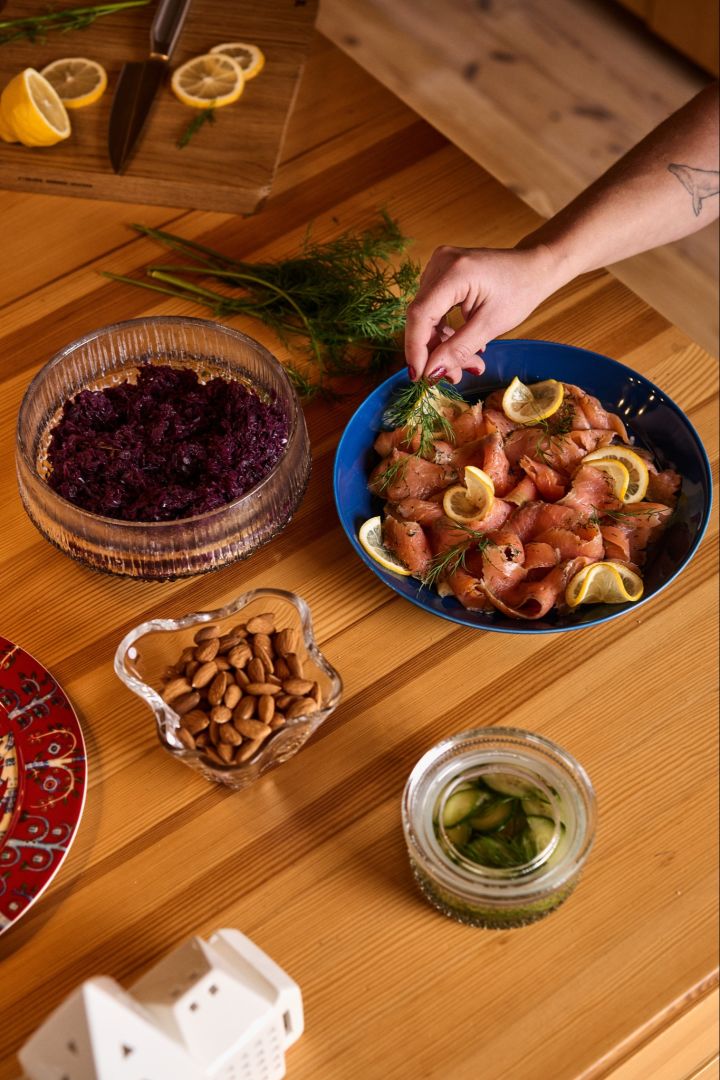 Schwedisches Weihnachtsessen: Eine Hand legt ein Stück Dill auf einen blauen Teller mit Lachs und Zitrone, ein traditionelles Gericht im Rahmen einer schwedischen Weihnachtsfeier. Daneben stehen Schalen mit Rotkohl und Mandeln.