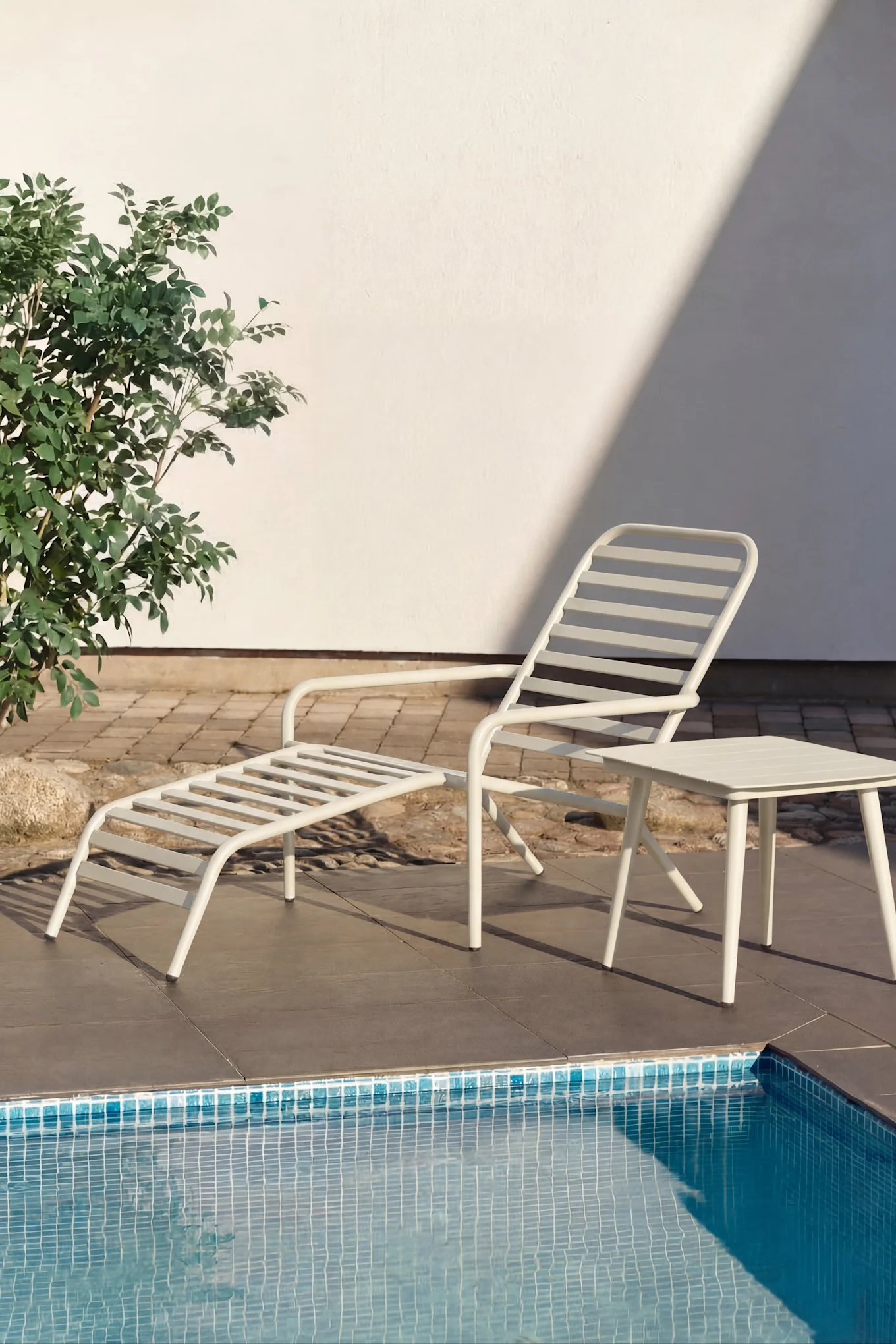 Weiße Sonnenliege und kleiner weißer Beistelltisch an einem Pool mit türkisfarbenem Wasser. Grüne Pflanzen und eine weiße Wand im Hintergrund.