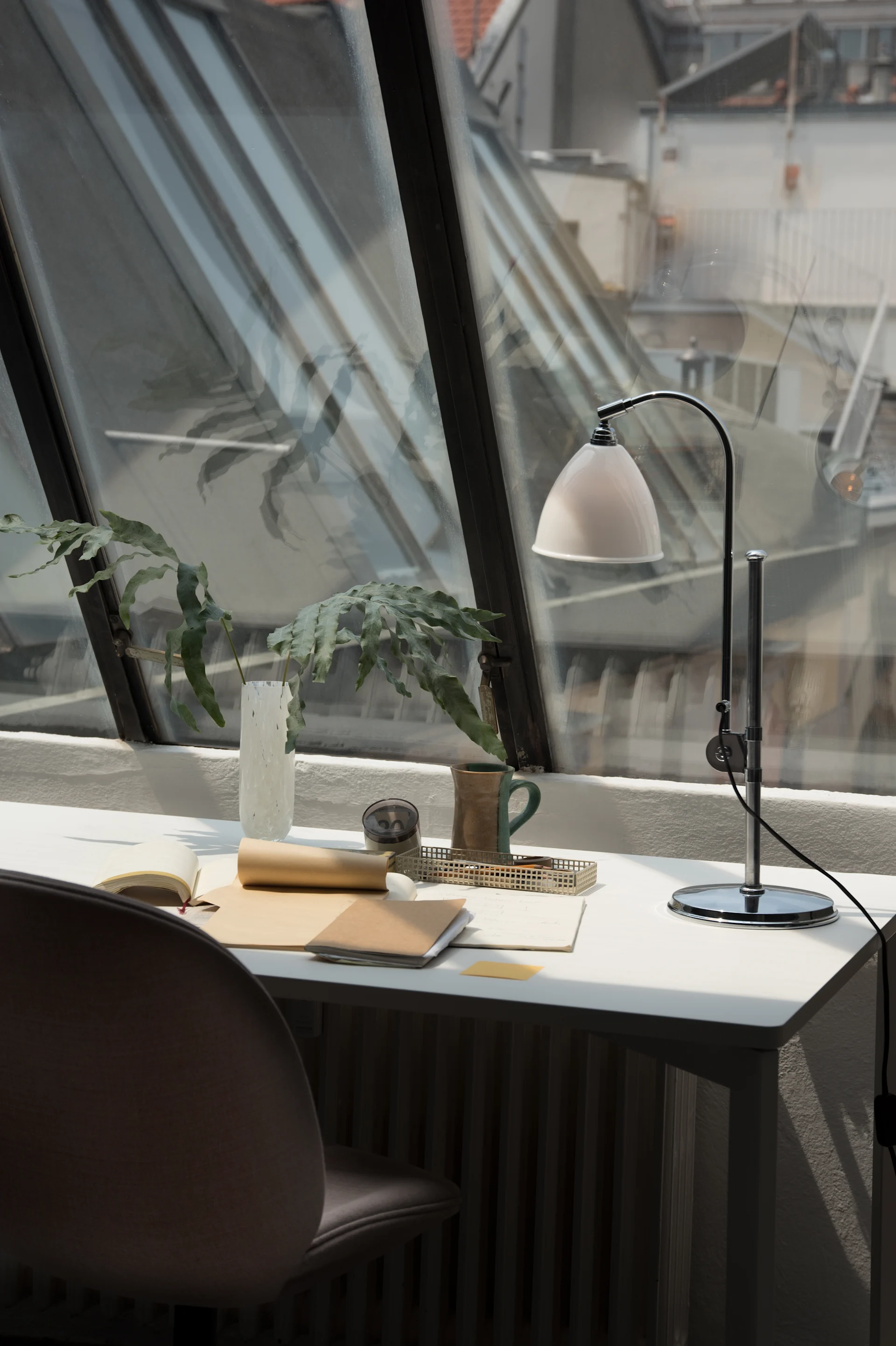 Moderner Schreibtischbereich an schrägem Fenster mit Blick auf Dächer, mit weißer Lampe, grüner Pflanze, Notizbüchern und zweifarbigem Becher.