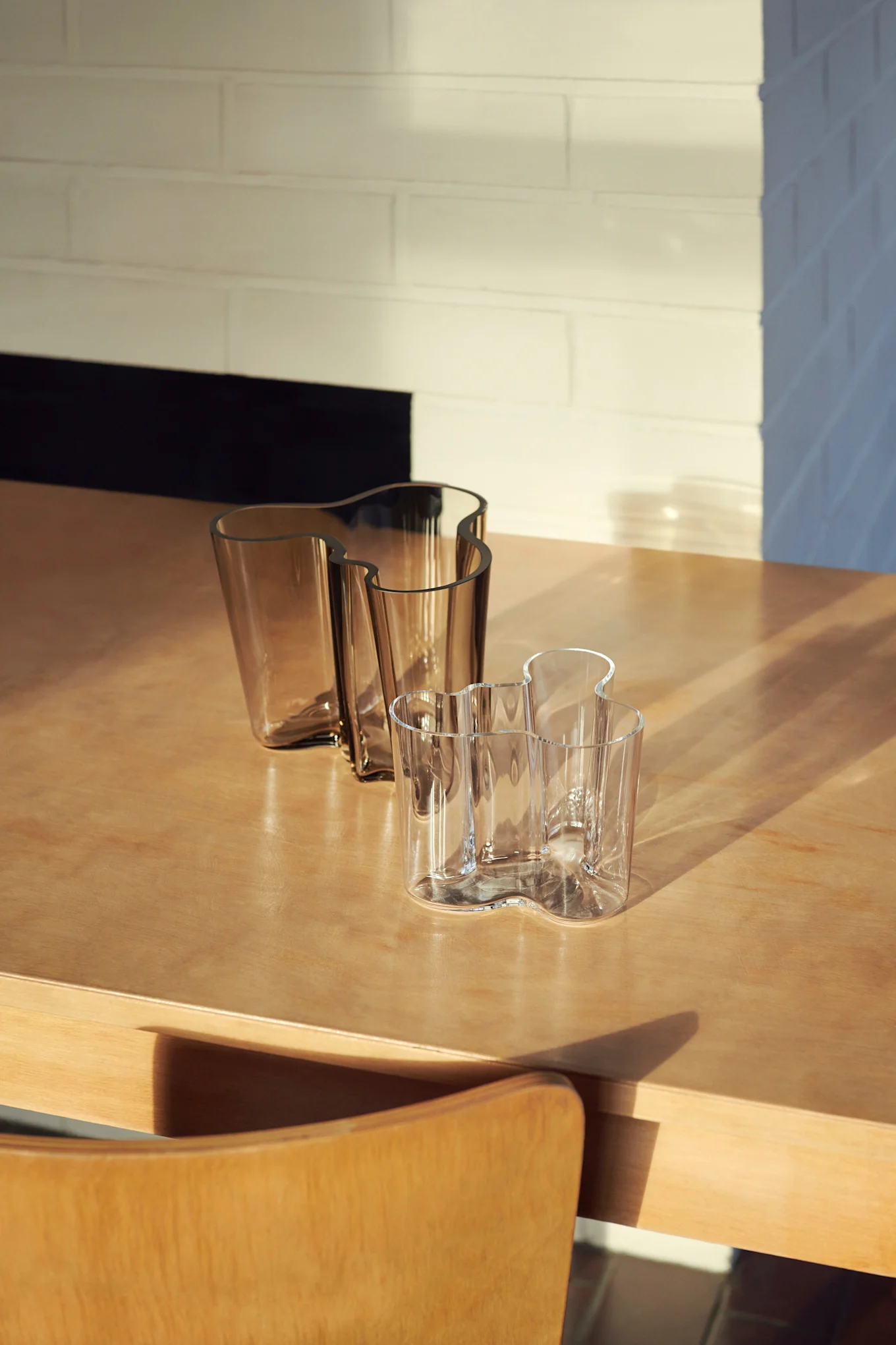 Drei gewellte Glasvasen auf einem Holztisch. Zwei sind bernsteinfarben, eine ist klar, mit Sonnenlicht und Schatten über der Szene.