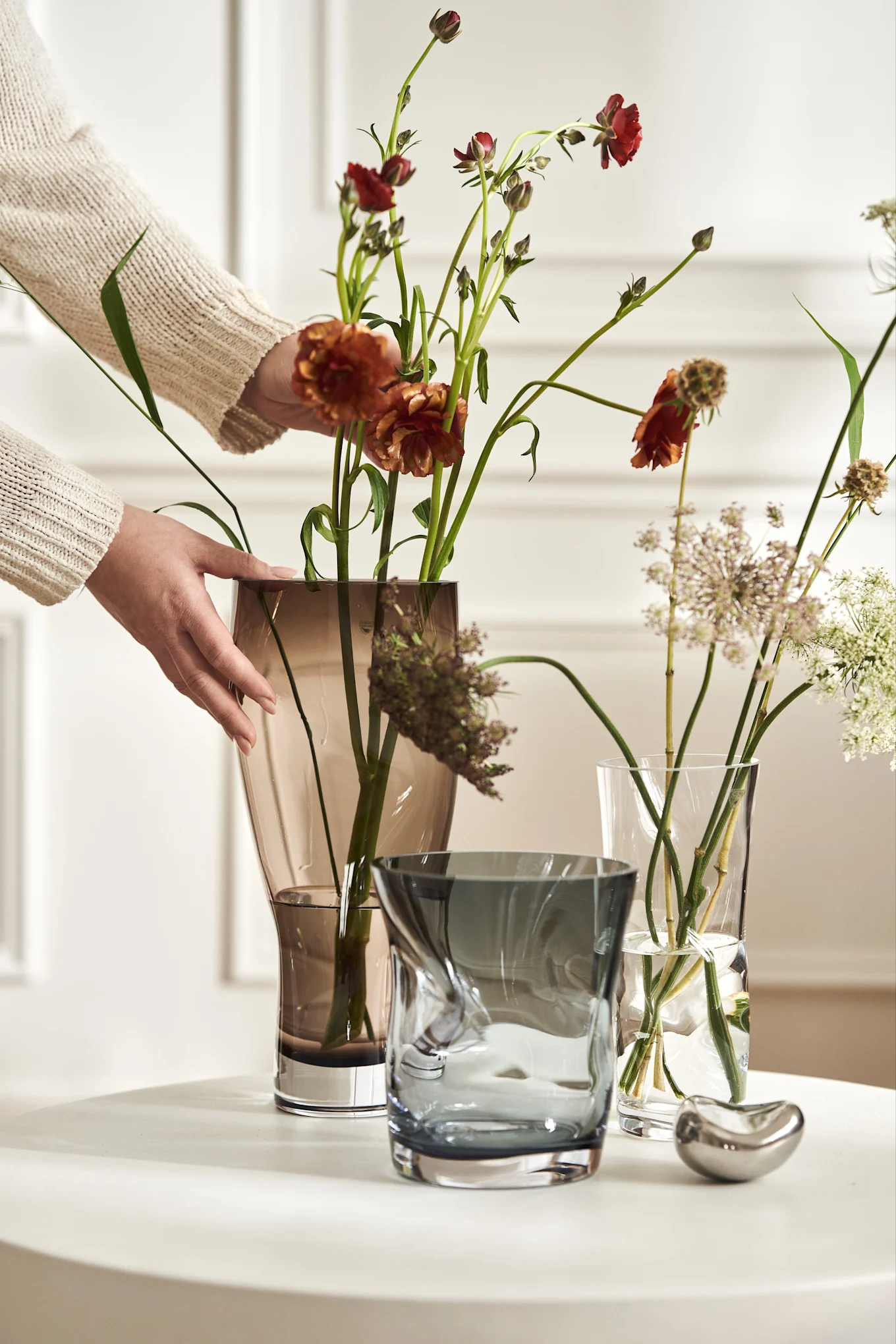 Hochzeitsgeschenk-Ideen: Drei Vasen aus der Squeeze-Kollektion von Orrefors stehen auf einem Tisch, während eine Hand die Blumen darin arrangiert.