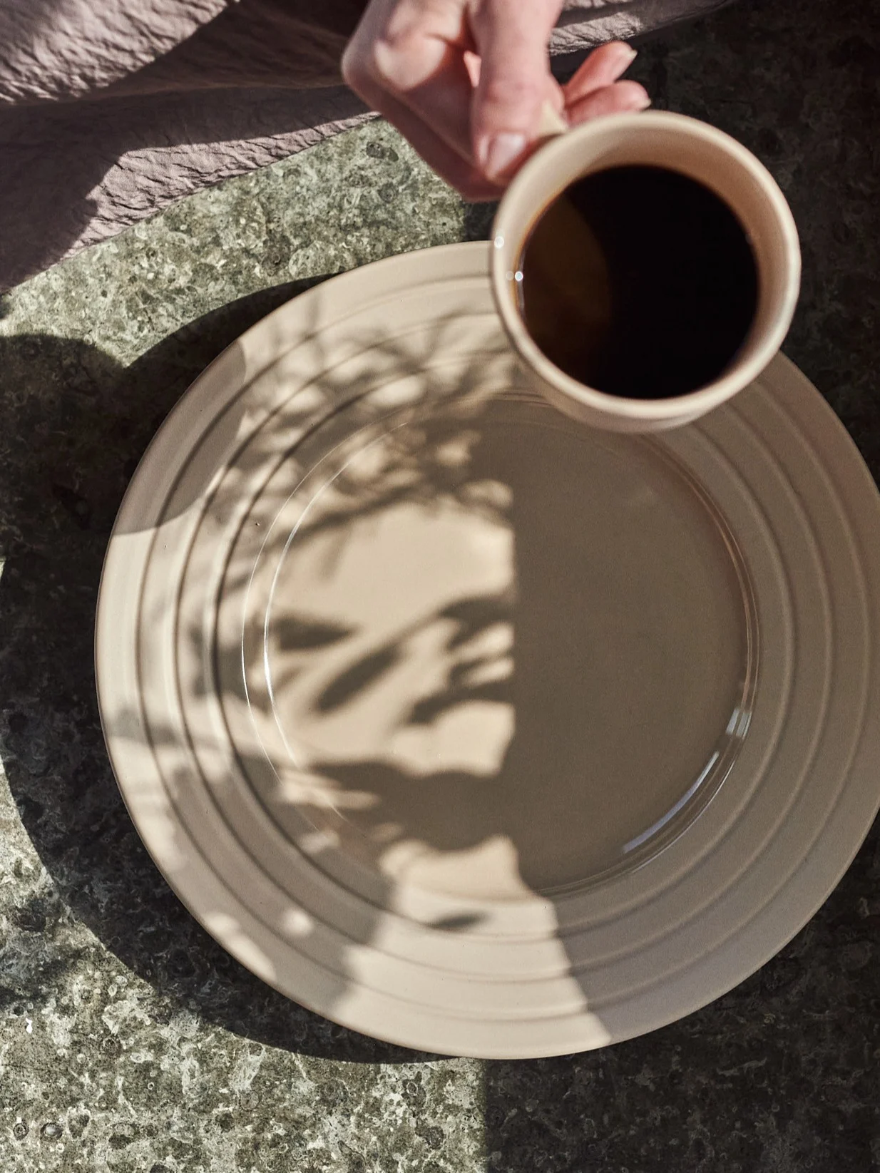 Hand hält Kaffeetasse über beigem Teller mit Blattschatten auf einem gesprenkelten Tisch.