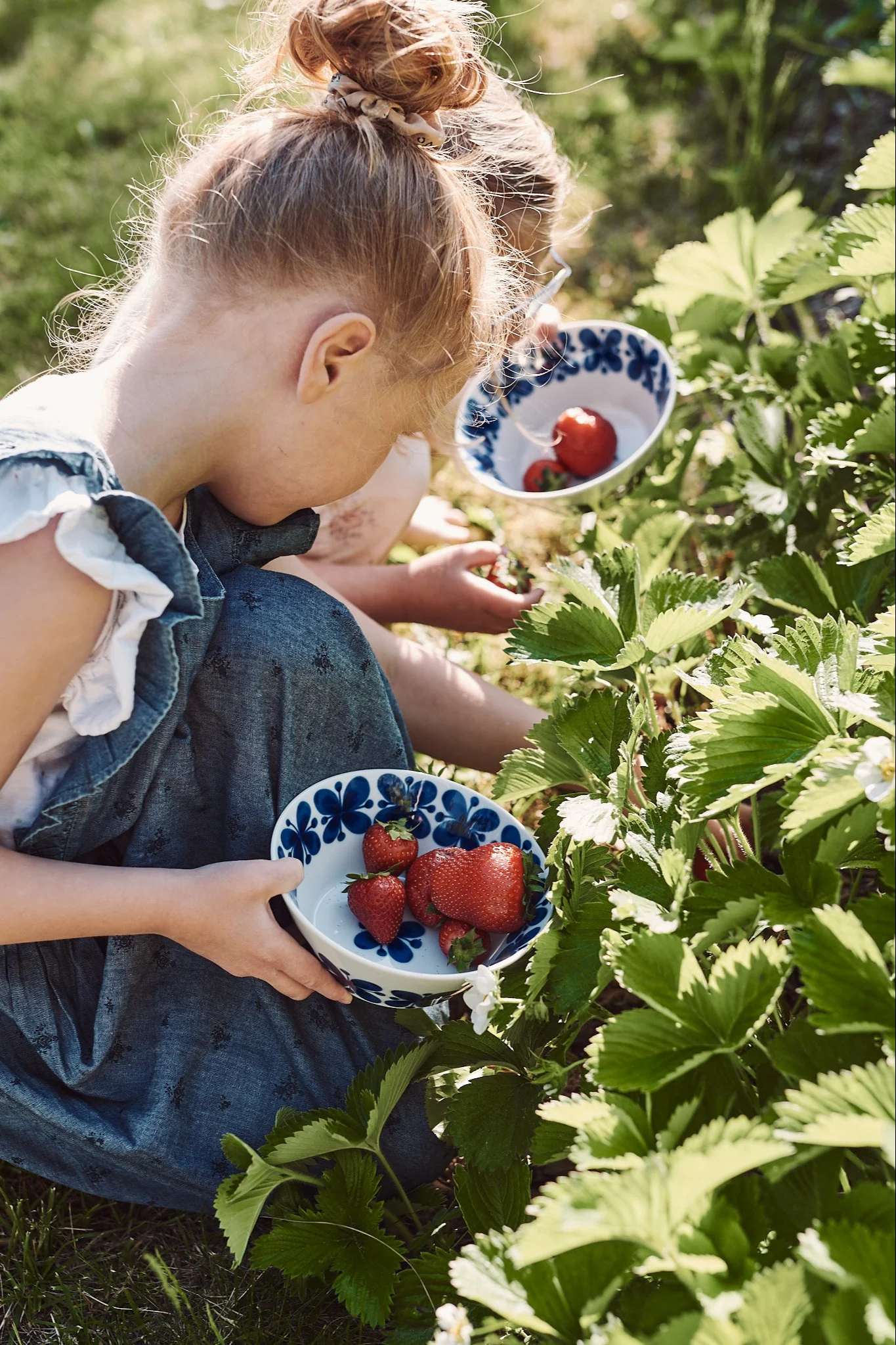 Summer Bucket List: Ein Mädchen kniet in einem Erdbeerfeld und sammelt Erdbeeren in die Mon Amie-Schale von Rörstrand.