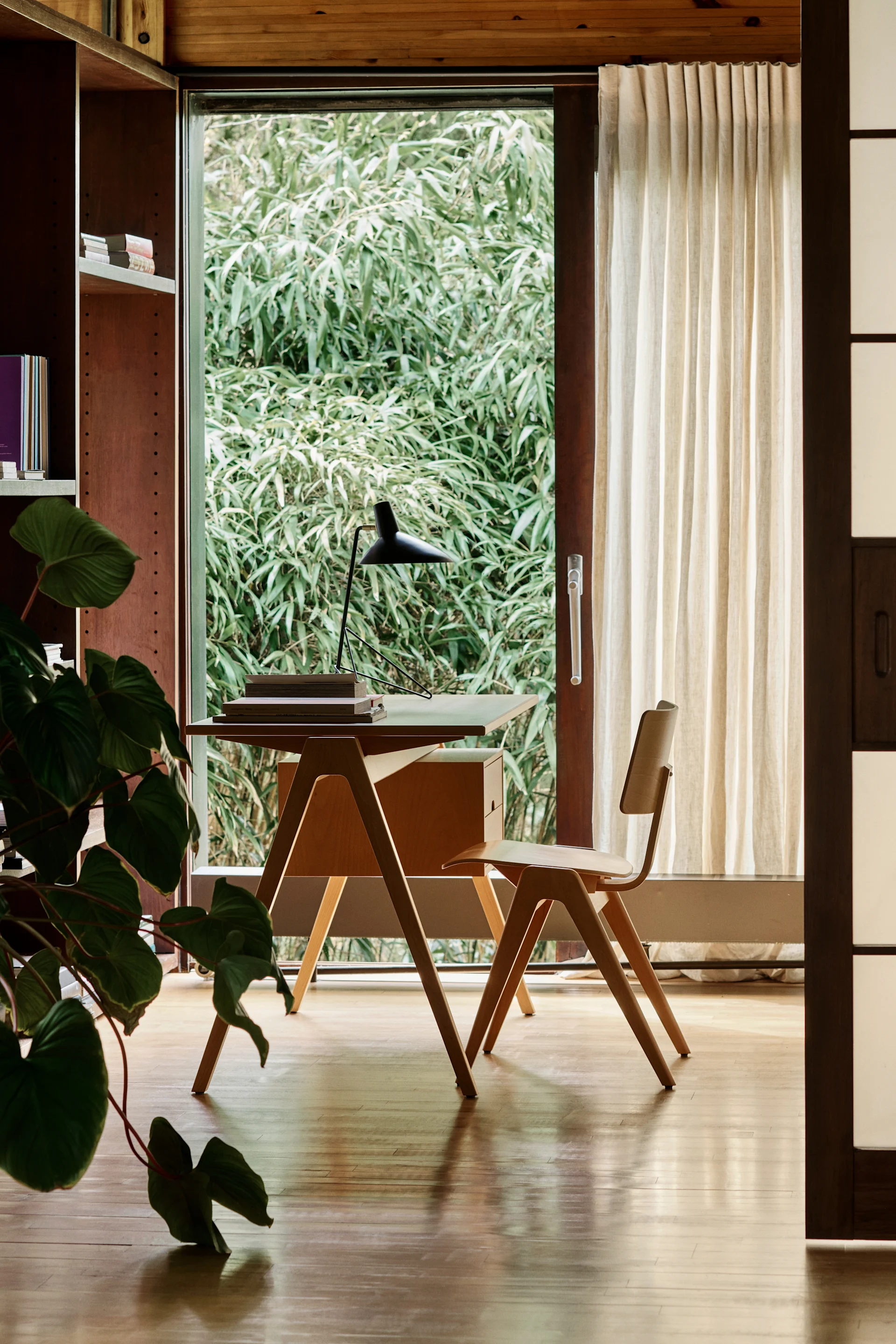 Minimalistisches Heimbüro mit Holztisch, Stuhl und Lampe, mit Blick auf grüne Außenbepflanzung.