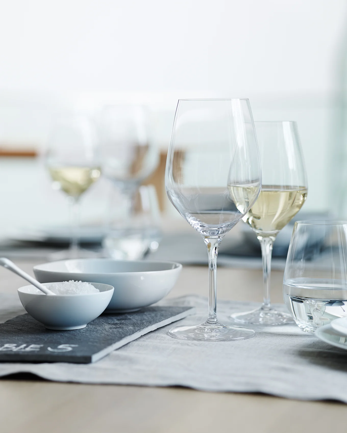 Ein gedeckter Esstisch mit klaren Wein- und Wassergläsern, einer Schale mit grobem Salz und einer leeren Schale auf einem grauen Tischset.