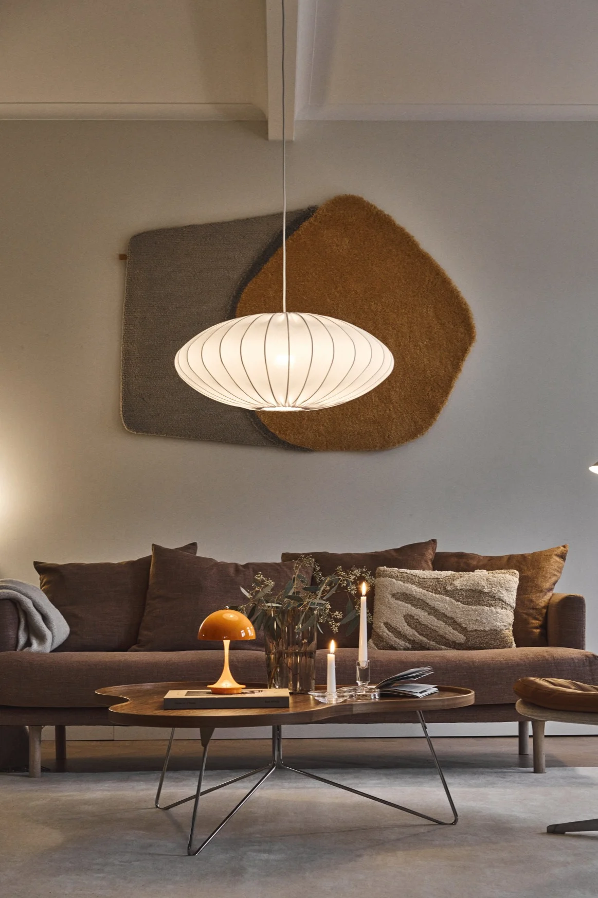 Gemütliches Wohnzimmer mit braunem Sofa, dunklem Couchtisch, Pilzlampe, Kerzen, weißer Pendelleuchte und Ledersessel.