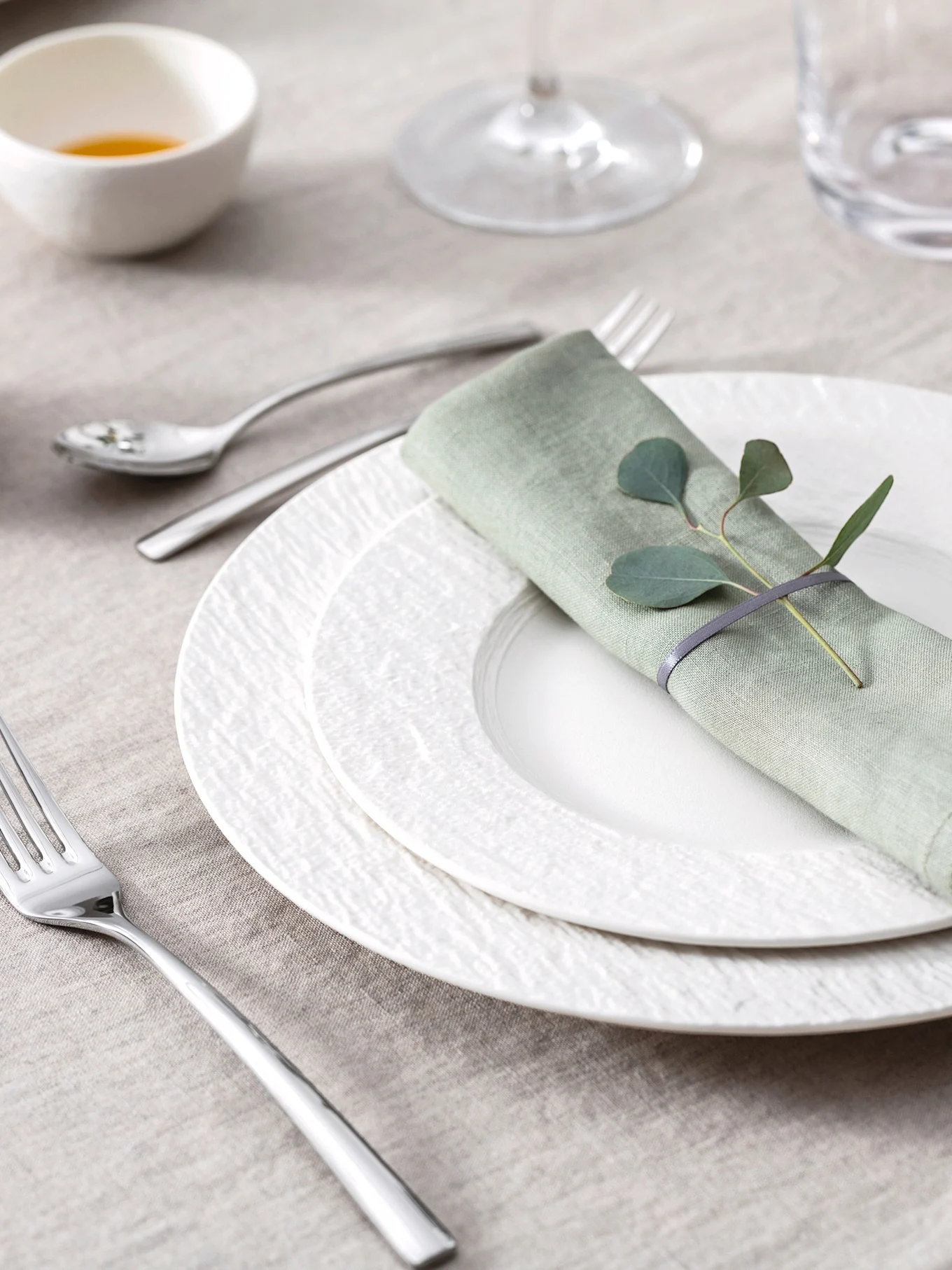 Elegante Tischdekoration mit hellgrüner Leinenserviette, Eukalyptusblättern, weißen strukturierten Tellern und Besteck auf einer beigen Tischdecke.