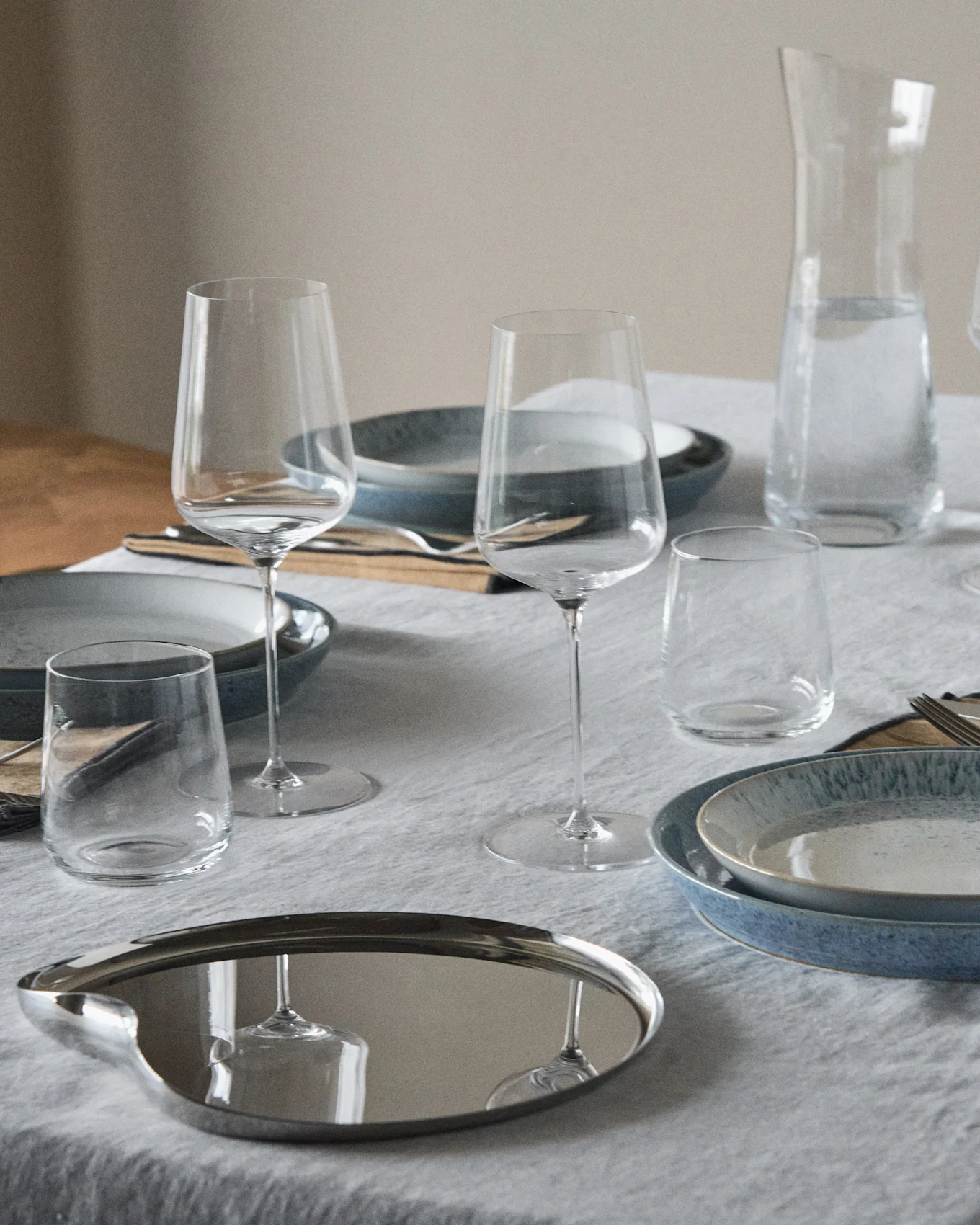 Ein gedeckter Tisch mit blauen und gesprenkelten Tellern, Weingläsern, Bechern, einer Wasserkaraffe und einem reflektierenden ovalen Tablett.