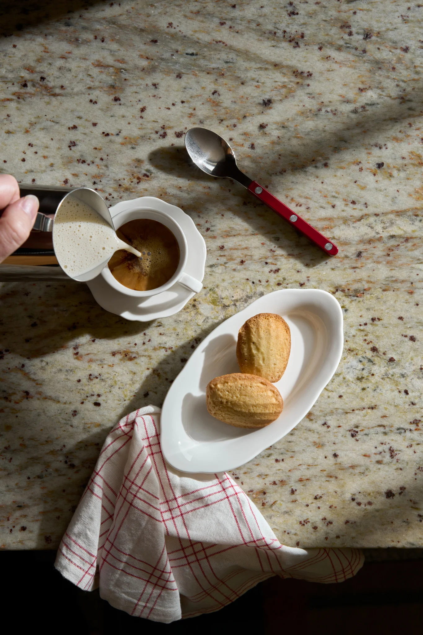Hand gießt aufgeschäumte Milch in Kaffee. Zwei Madeleines auf einem weißen Teller und ein Löffel mit rotem Griff liegen auf einer Granitplatte.
