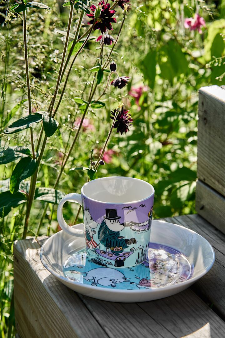 Die Sommer-Mumin-Tasse 2025 "Beach Day" steht mit dem dazugehörigen Teller auf einer Treppenstufe, im Hintergrund ist eine wilde Wiese zu sehen.