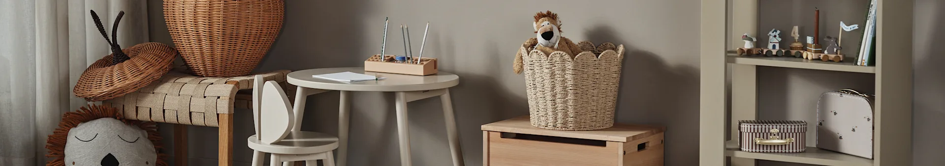 Kinderzimmer mit Löwenkuscheltier in einem Weidenkorb, kleinem Holztisch mit Stiften und einem Bücherregal mit Spielzeug und Büchern.