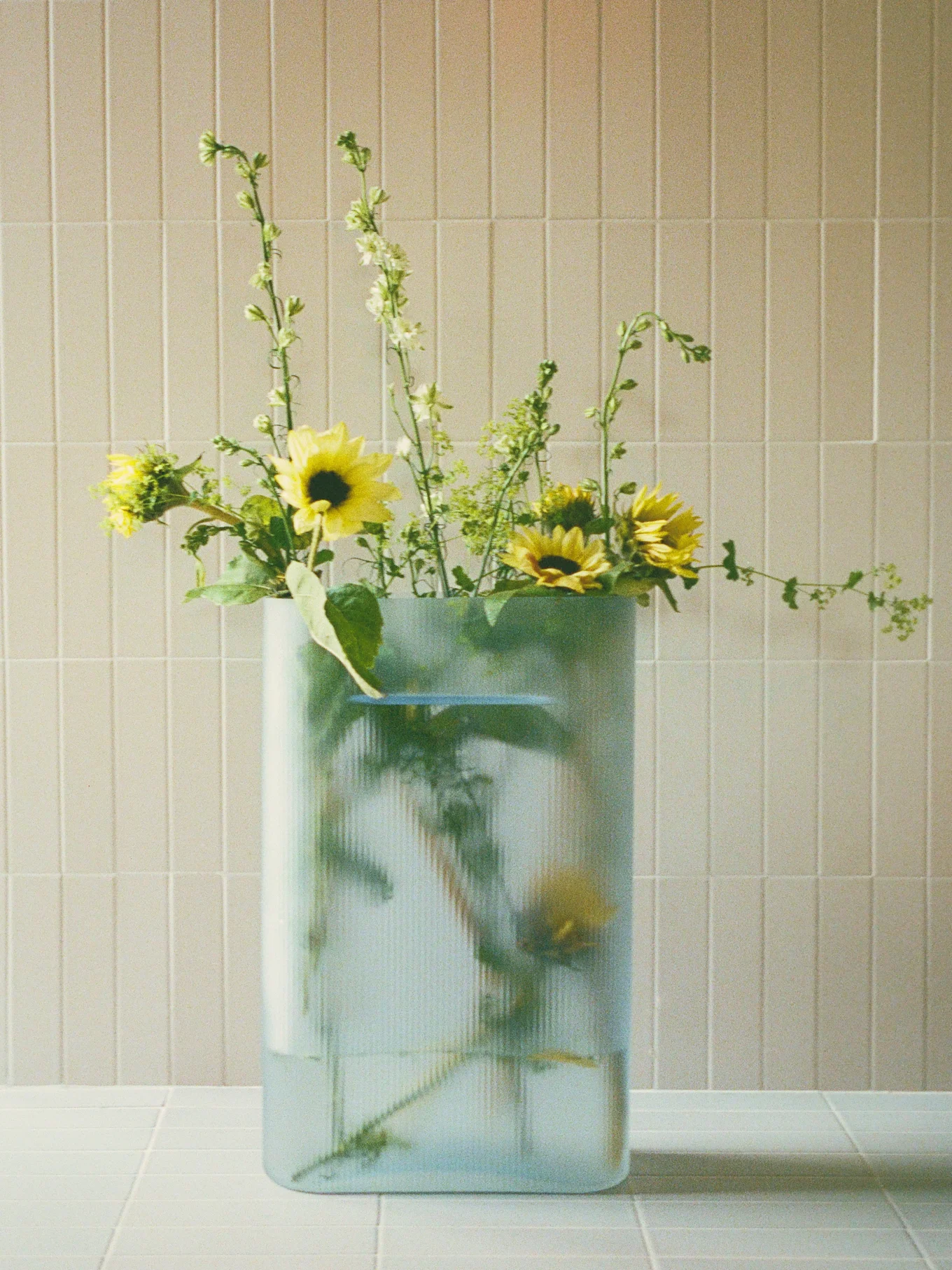 Strauß aus gelben Sonnenblumen und grünen, stacheligen Blumen in einer klaren, gerippten rechteckigen Vase auf einer gefliesten Oberfläche.