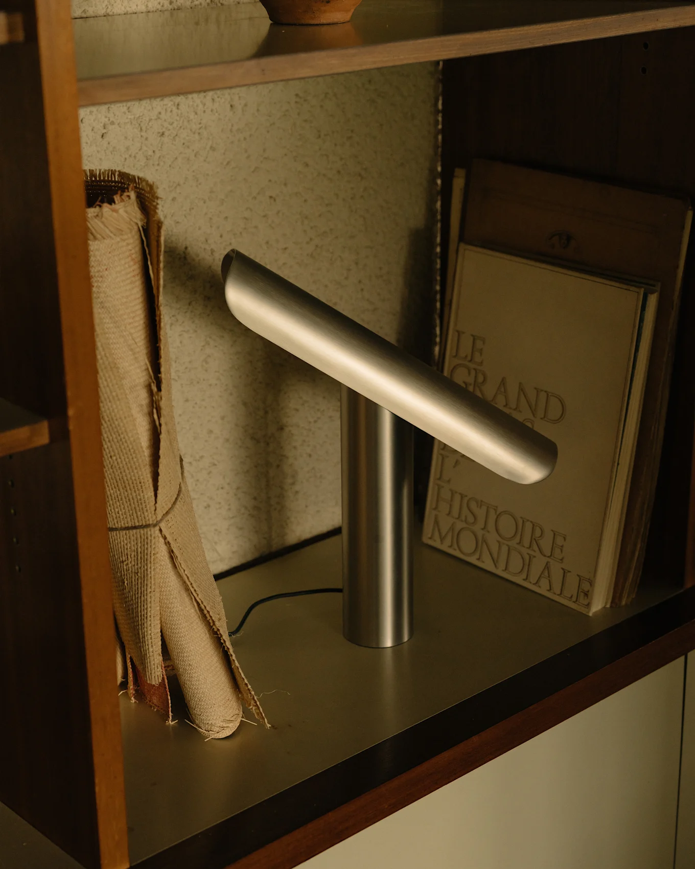 Moderne silberne Schreibtischlampe mit zylindrischem Sockel und einer abgewinkelten horizontalen Lichtleiste auf einem Regal.