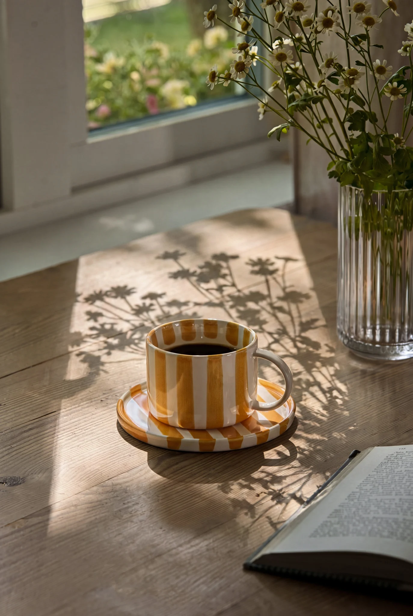 Orange-weiß gestreifte Kaffeetasse mit schwarzem Kaffee, eine Vase mit weißen Blumen und ein offenes Buch auf einem sonnenbeschienenen Holztisch.
