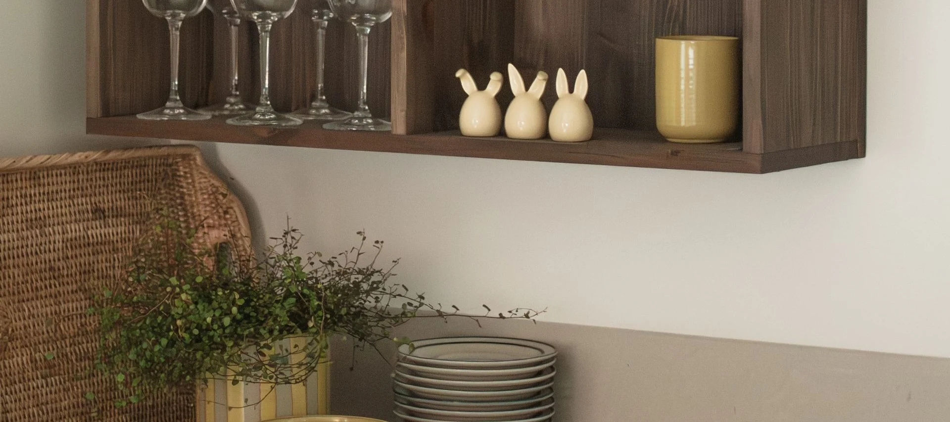 Ein Holzregal mit Gläsern, drei Hasenfiguren und einem gelben Becher, darunter ein Weidenkorb und Pflanzen.