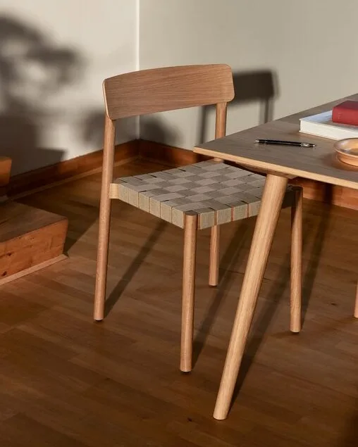 Ein heller Holzstuhl mit geflochtener karierter Sitzfläche neben einem Tisch mit Büchern und einem Stift.