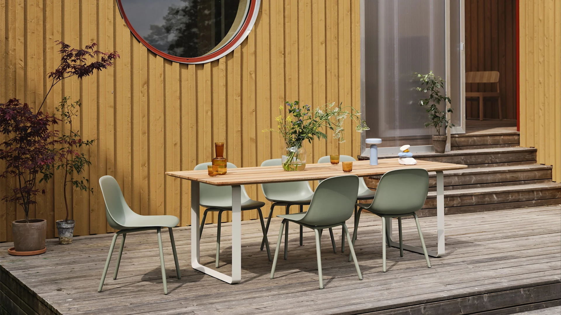 Gartenmöbel Trends 2025: Die salbeigrünen Fiber Outdoor-Stühle von Muuto stehen auf einer großen Terrasse und einen Tisch herum, im Hintergrund ist ein gelbes Holzhaus zu sehen.