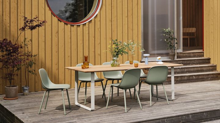 Gartenmöbel Trends 2025: Die salbeigrünen Fiber Outdoor-Stühle von Muuto stehen auf einer großen Terrasse und einen Tisch herum, im Hintergrund ist ein gelbes Holzhaus zu sehen.