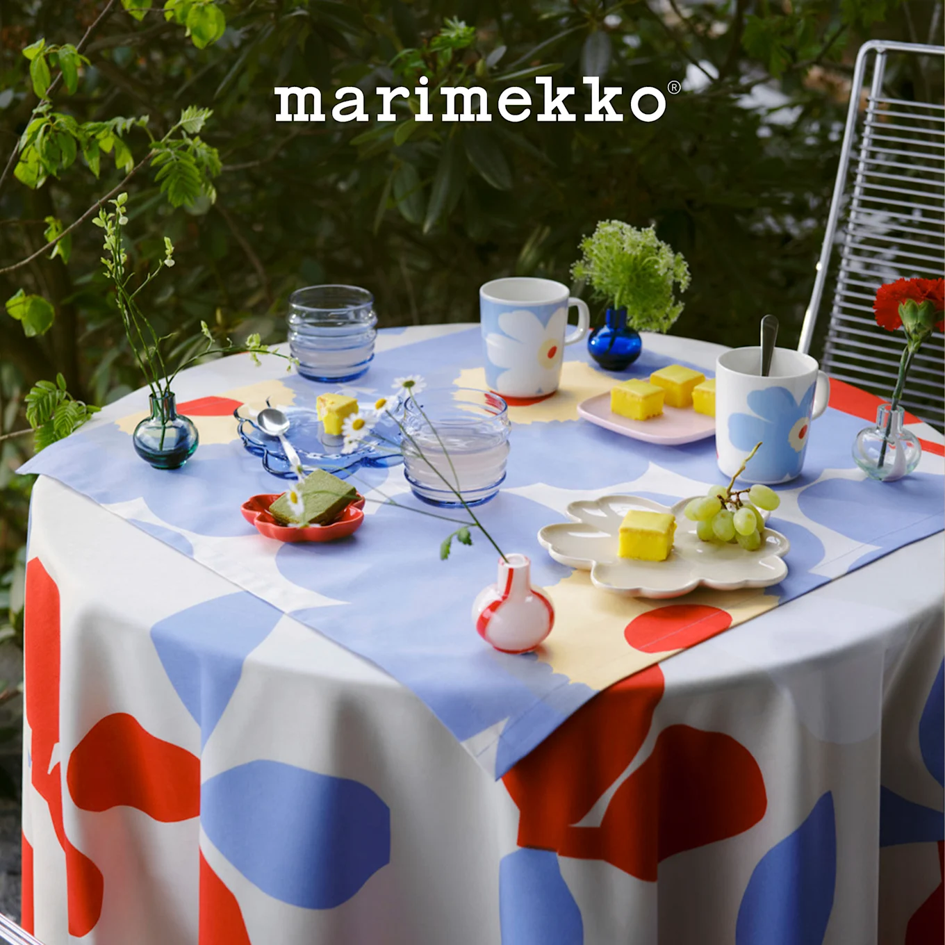 Outdoor-Tisch mit Marimekko rot, blau, weiß gemusterter Tischdecke, gedeckt mit Blumenbechern, Desserts und kleinen Pflanzen. Marimekko Logo.