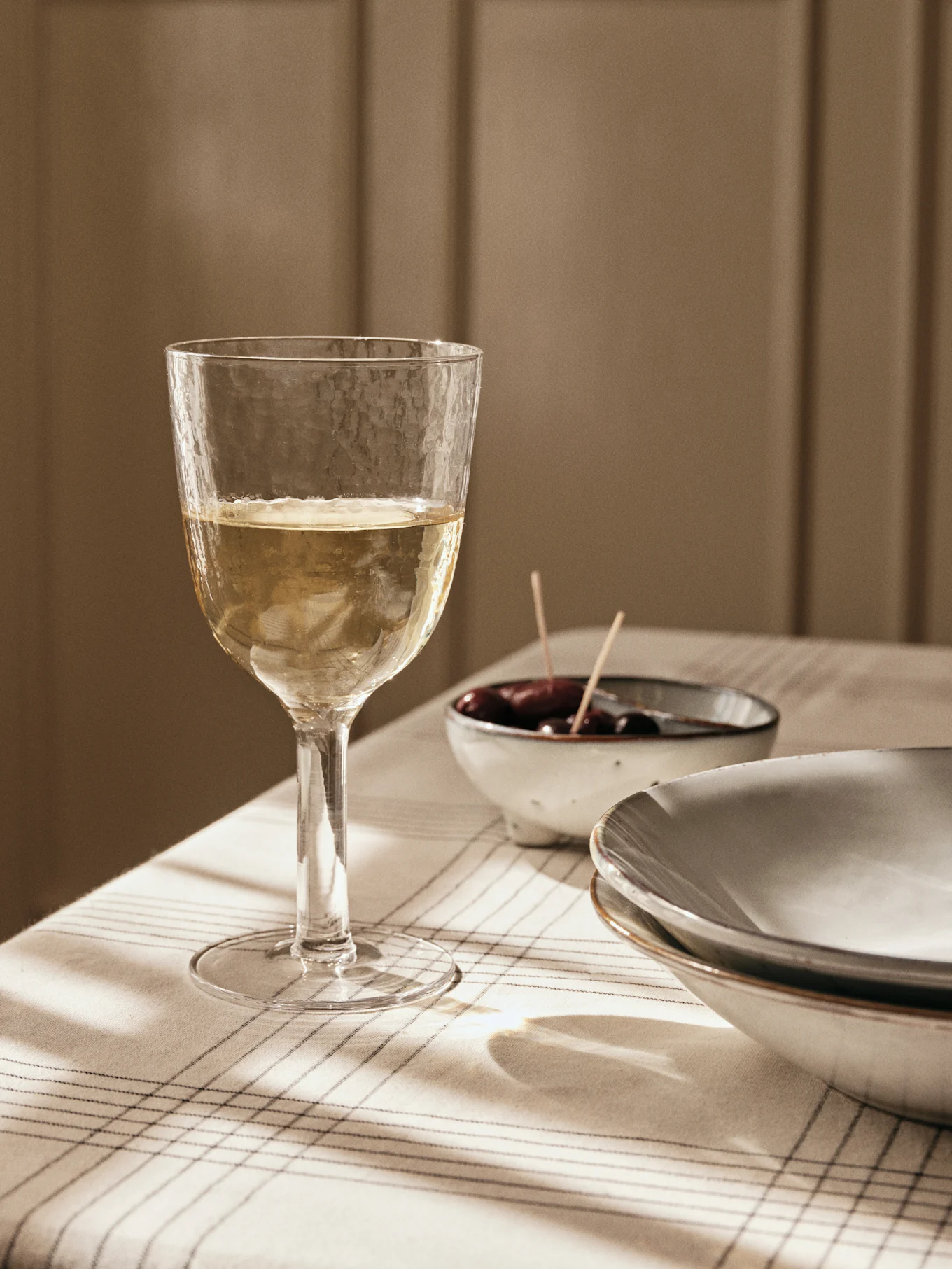 Ein strukturiertes Glas Weißwein auf einer gestreiften Tischdecke, mit Oliven und Tellern im Hintergrund, gebadet in Fensterlicht.