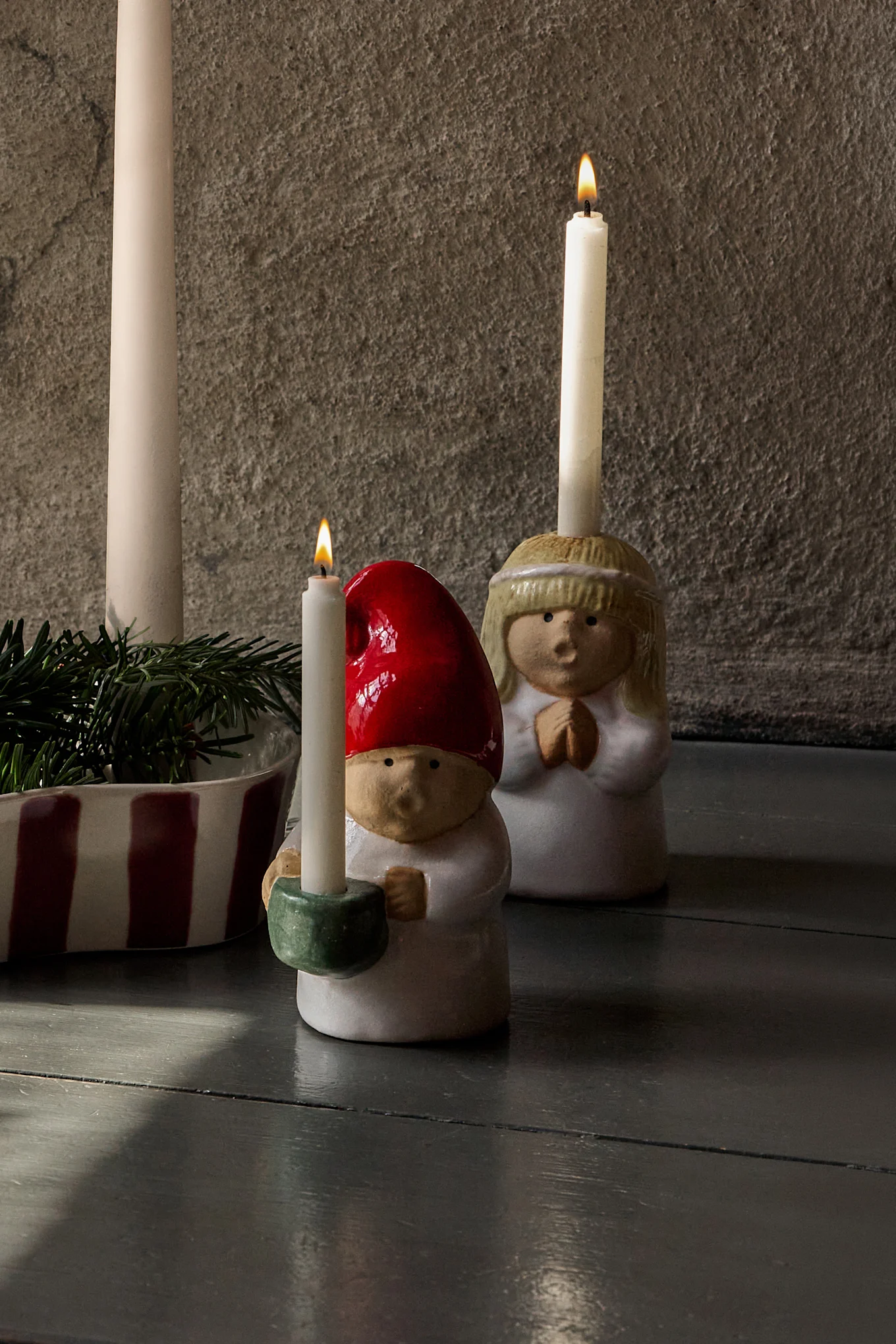 Adventsarrangement: vier unangezündete Kerzen in gestreiftem Halter mit Tannengrün, flankiert von Keramikfiguren (Engel und Wichtel) mit angezündeten Kerzen.