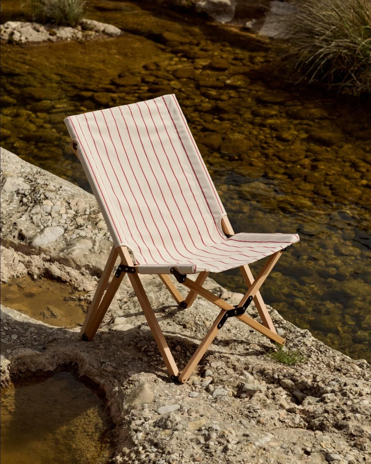 Klappstuhl mit weiß-rot gestreiftem Stoff auf einem felsigen Ufer neben einem Fluss.