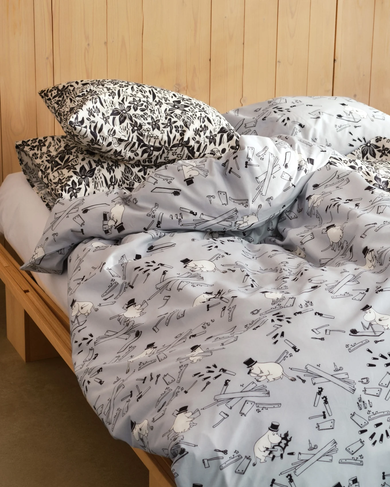 Bett mit Kissen in schwarz-weißem Blumendruck und einer hellblauen Bettdecke mit Zeichentrickfiguren, Werkzeugen und Holzbrettern. Bettrahmen aus Holz.