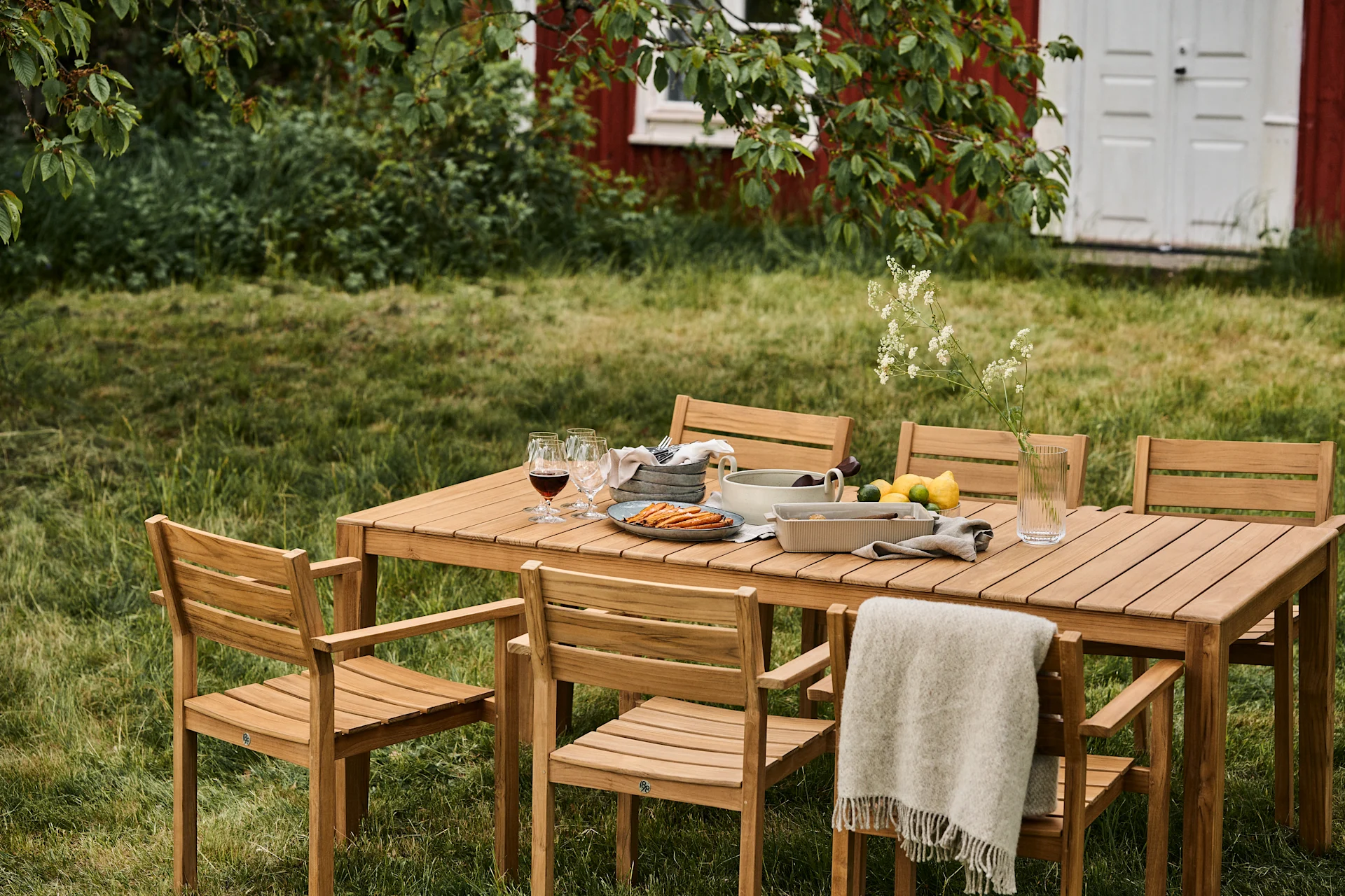 Outdoor-Essgruppe mit Holztisch und sechs Stühlen auf einem Rasen, gedeckt mit Speisen, Getränken und einer Decke, mit einem roten Haus im Hintergrund.