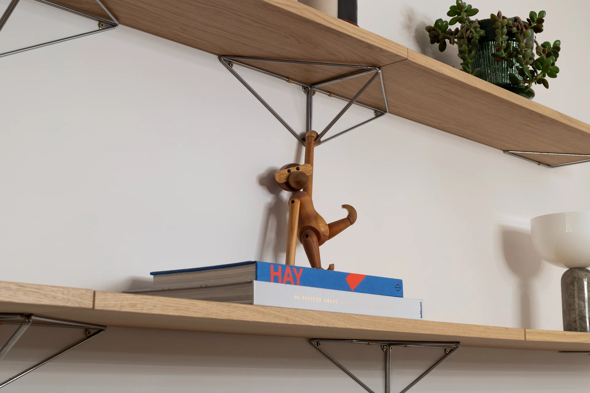 Zwei Holzregale an weißer Wand. Oben: gestreifte Kerzen, Topfpflanze. Unten: Bücher, Holzaffe, weiße Lampe.