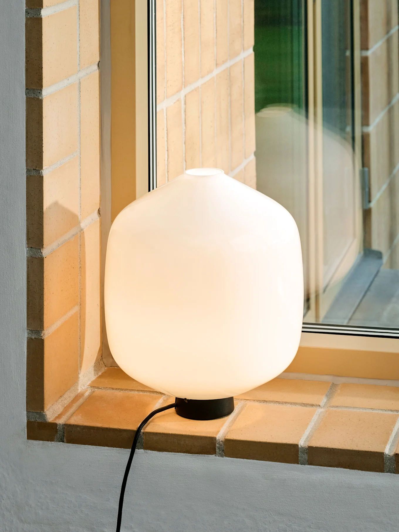 Eine leuchtende, abgerundete weiße Tischlampe mit schwarzem Fuß und Kabel auf einer gemauerten Fensterbank.