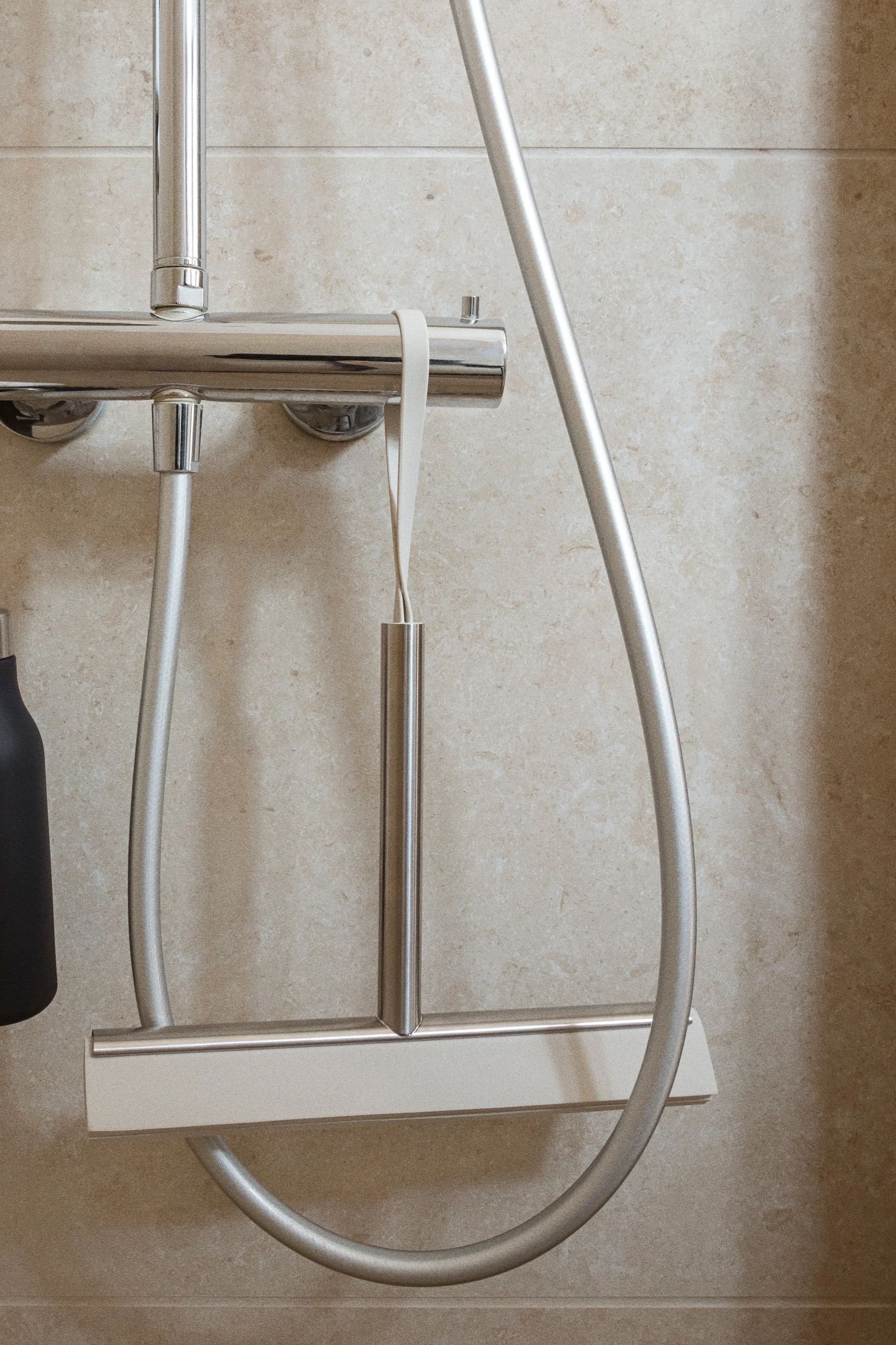 Ein Duschabzieher aus gebürstetem Metall mit heller Klinge hängt an einer verchromten Duscharmatur in einer gefliesten Dusche.