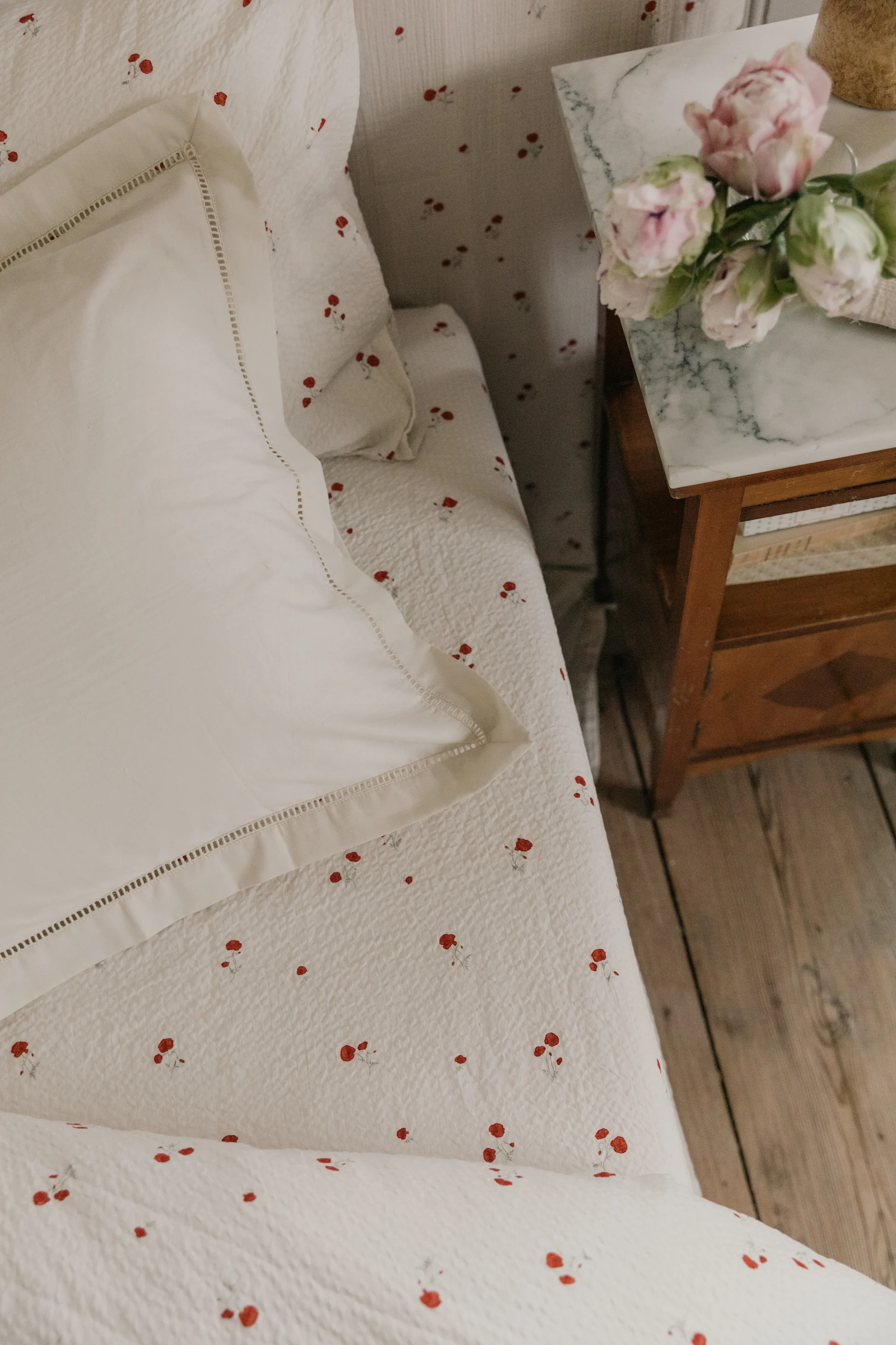 Weiße Bettwäsche mit rotem Blumenmuster, ein weißes Kissen und rosa Blumen auf einem Marmornachttisch.