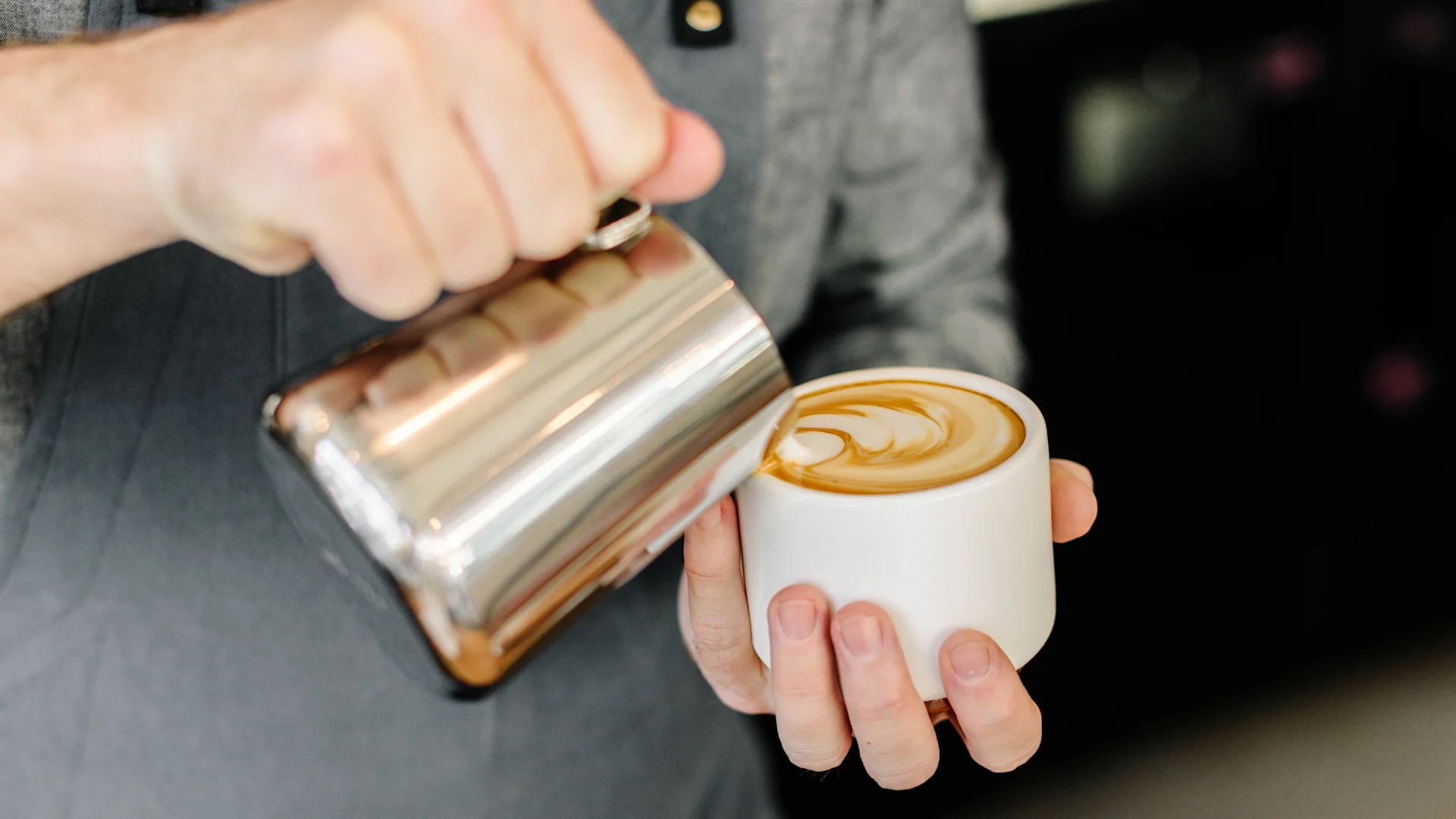 Barista gießt Milch aus einer silbernen Kanne in eine weiße Tasse und kreiert Latte Art.