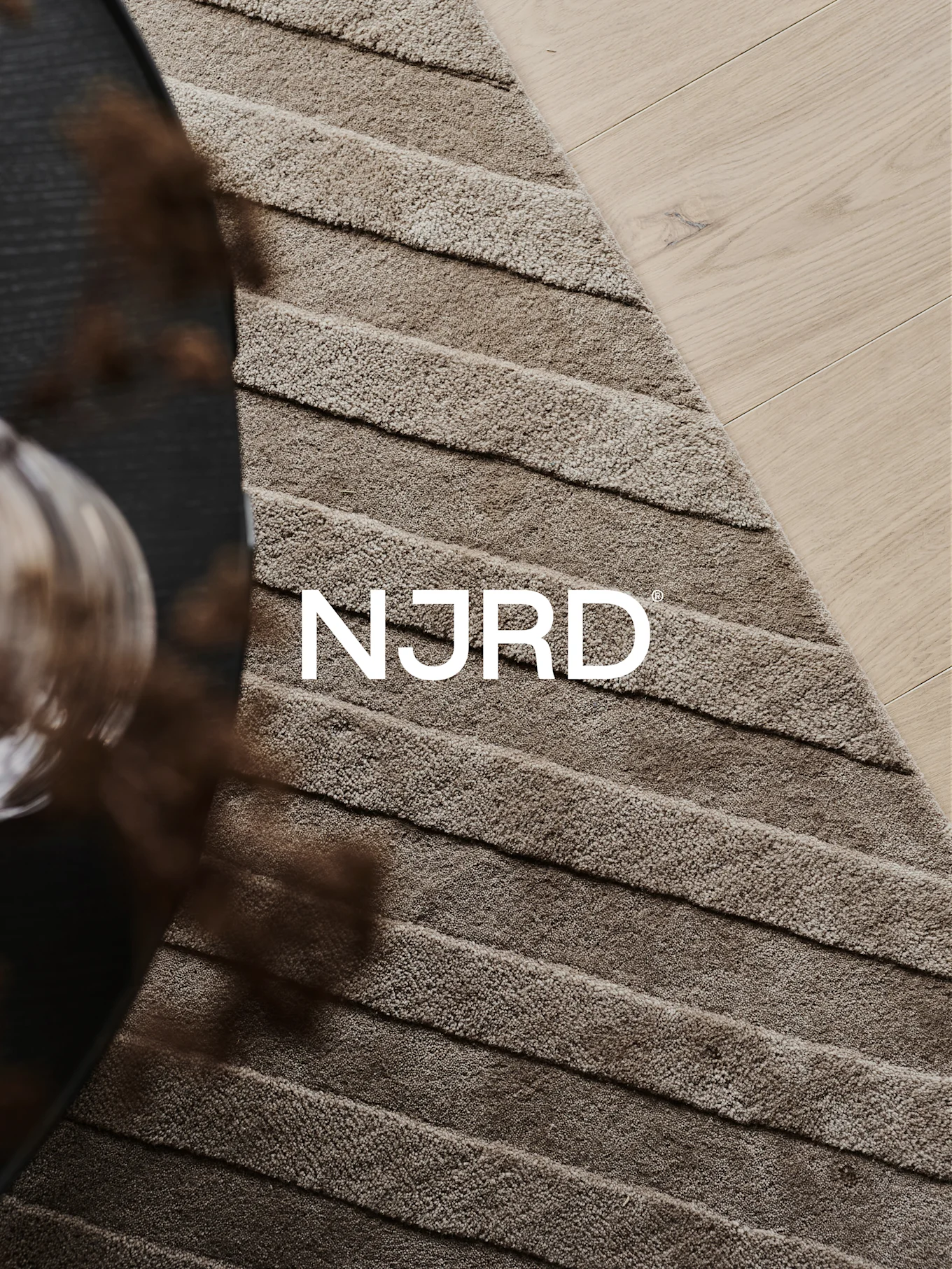 Nahaufnahme eines hellbraunen Hochflorteppichs mit horizontalen Rillen, teilweise auf hellem Holzboden, mit dem weißen 'NJRD'-Logo.