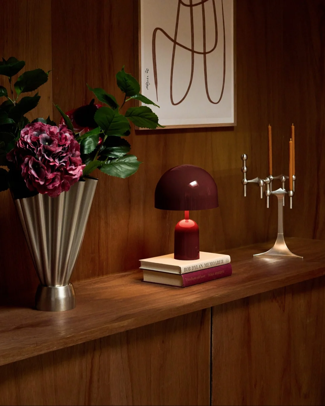 Gemütliches Holzregal mit silberner Rillen-Vase, dunkelroten Hortensien, roter Lampe auf Büchern und silbernem Kerzenständer.