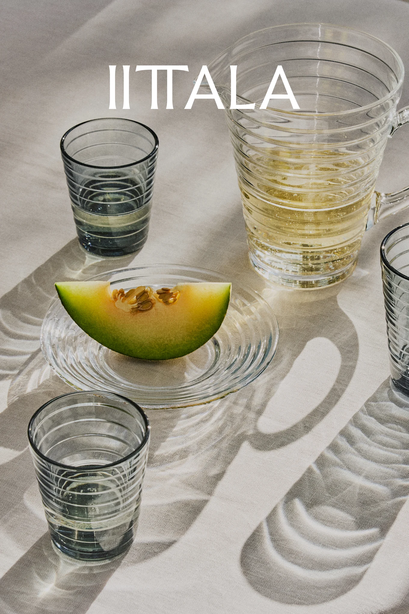IITTALA Logo über Gläsern, einem Krug mit Sprudelgetränk und einer Melonenscheibe auf Teller, auf hellem Stoff.