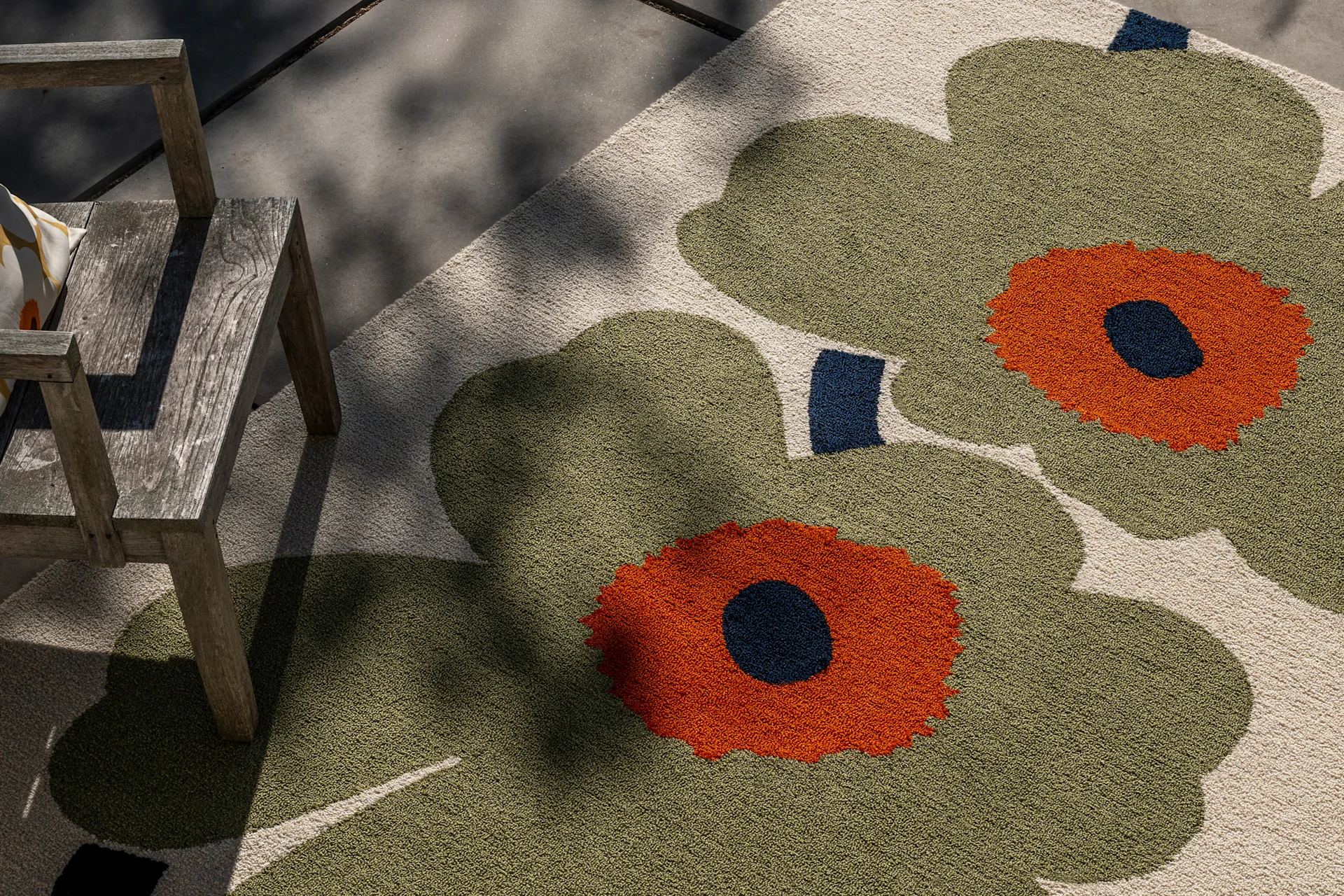 Outdoor-Teppich mit großem olivgrünem, orangem und dunkelblauem Blumenmuster, neben einem rustikalen Holzstuhl im gefleckten Sonnenlicht.