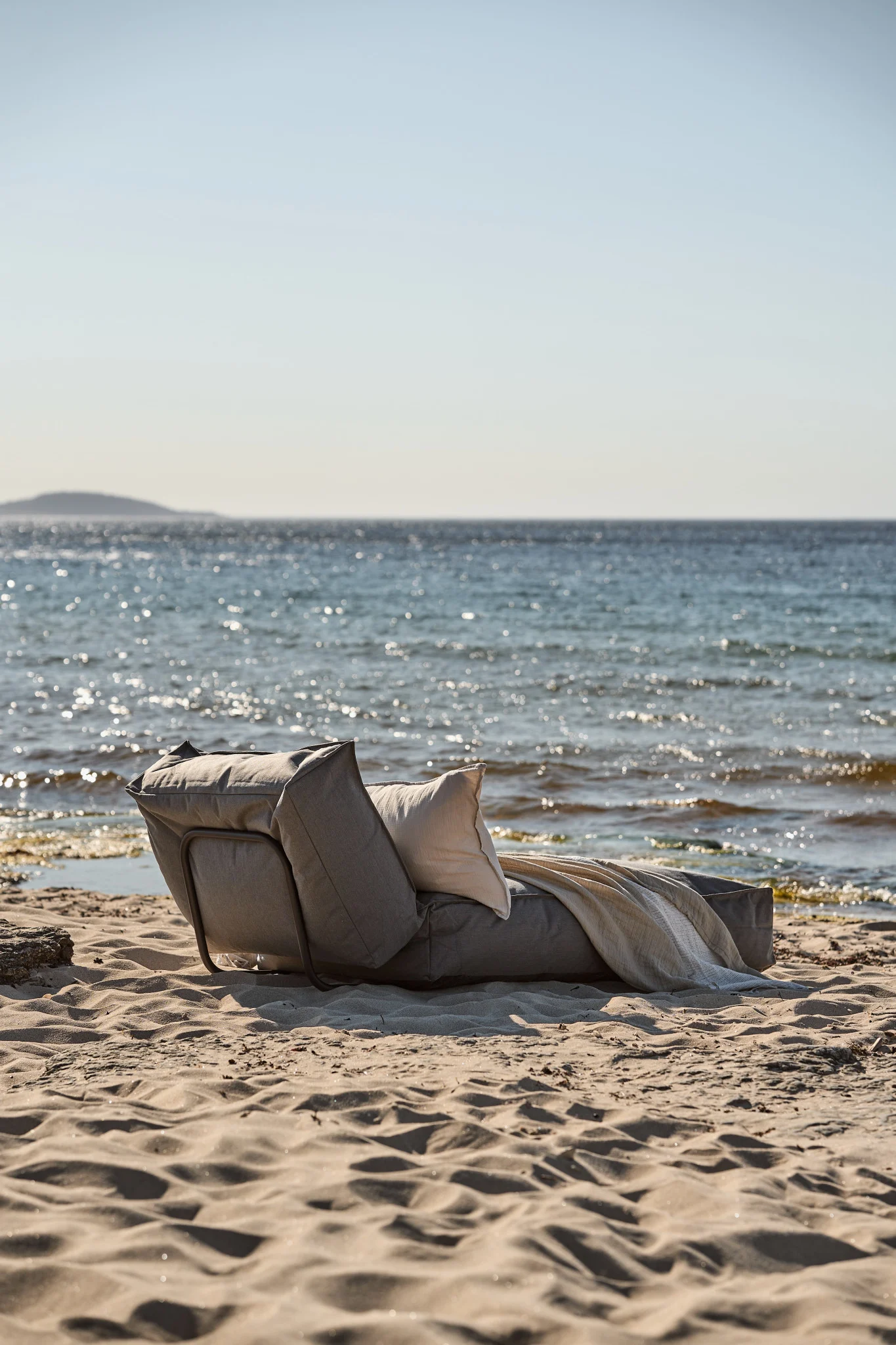 Grauer Liegestuhl mit beigem Kissen und heller Decke am Sandstrand am Meer.