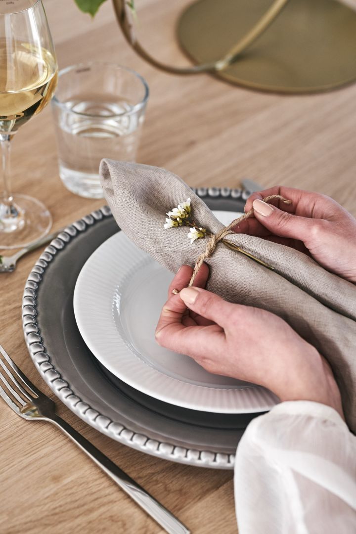 Beigefarbene Leinenserviette von Scandi Living, gefaltet wie ein Knoten und gebunden mit Schnur und Blume auf einem festlich gedeckten Tisch. 