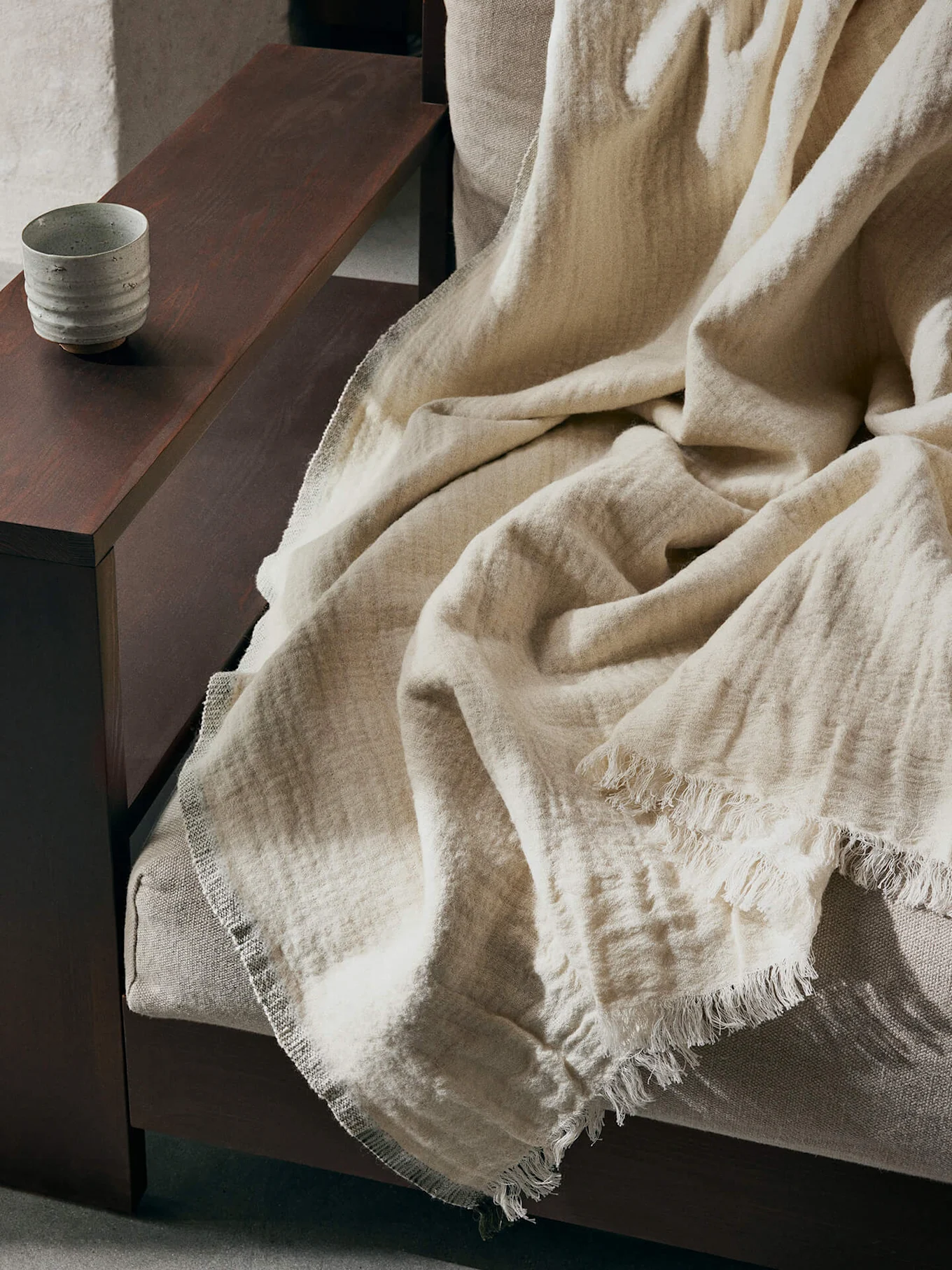 Eine hellbeige, texturierte Decke mit ausgefransten Rändern liegt über einem Sofa, mit einer Keramiktasse auf einer dunklen Holzarmlehne.