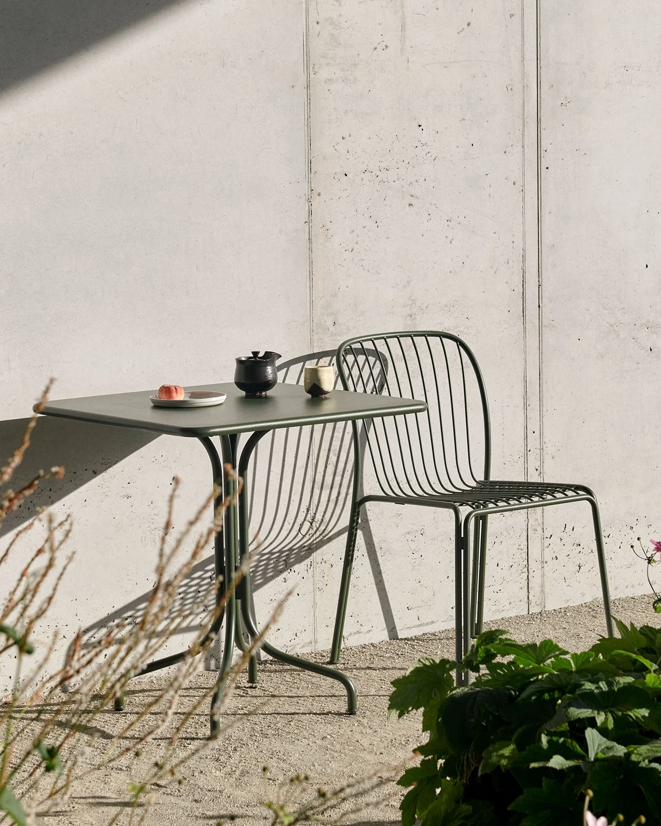 Ein dunkelgrüner Gartentisch mit einem Pfirsich, Teekanne und Tasse sowie ein passender Lattenstuhl vor einer Betonwand mit starken Schatten.