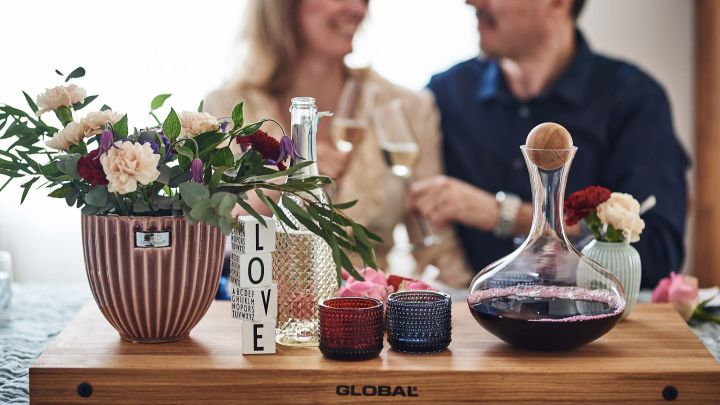 Der Tisch ist für ein romantisches Abendessen mit einem großen Schneidebrett von Global in der Mitte des Tisches eingerichtet, auf dem eine Weinkaraffe steht. 
