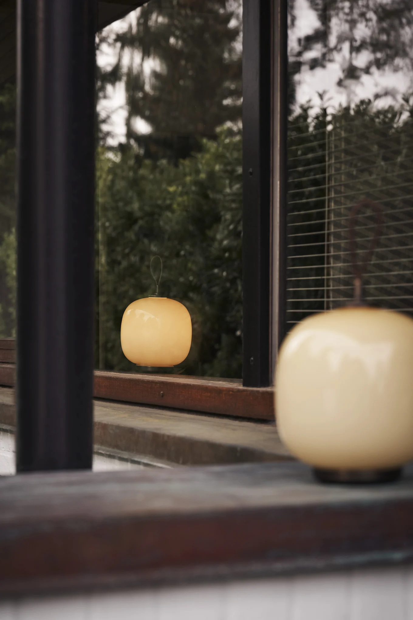 Zwei helle, runde Lampen, eine unscharf im Vordergrund und eine in einem Fenster mit grünem Laub gespiegelt.