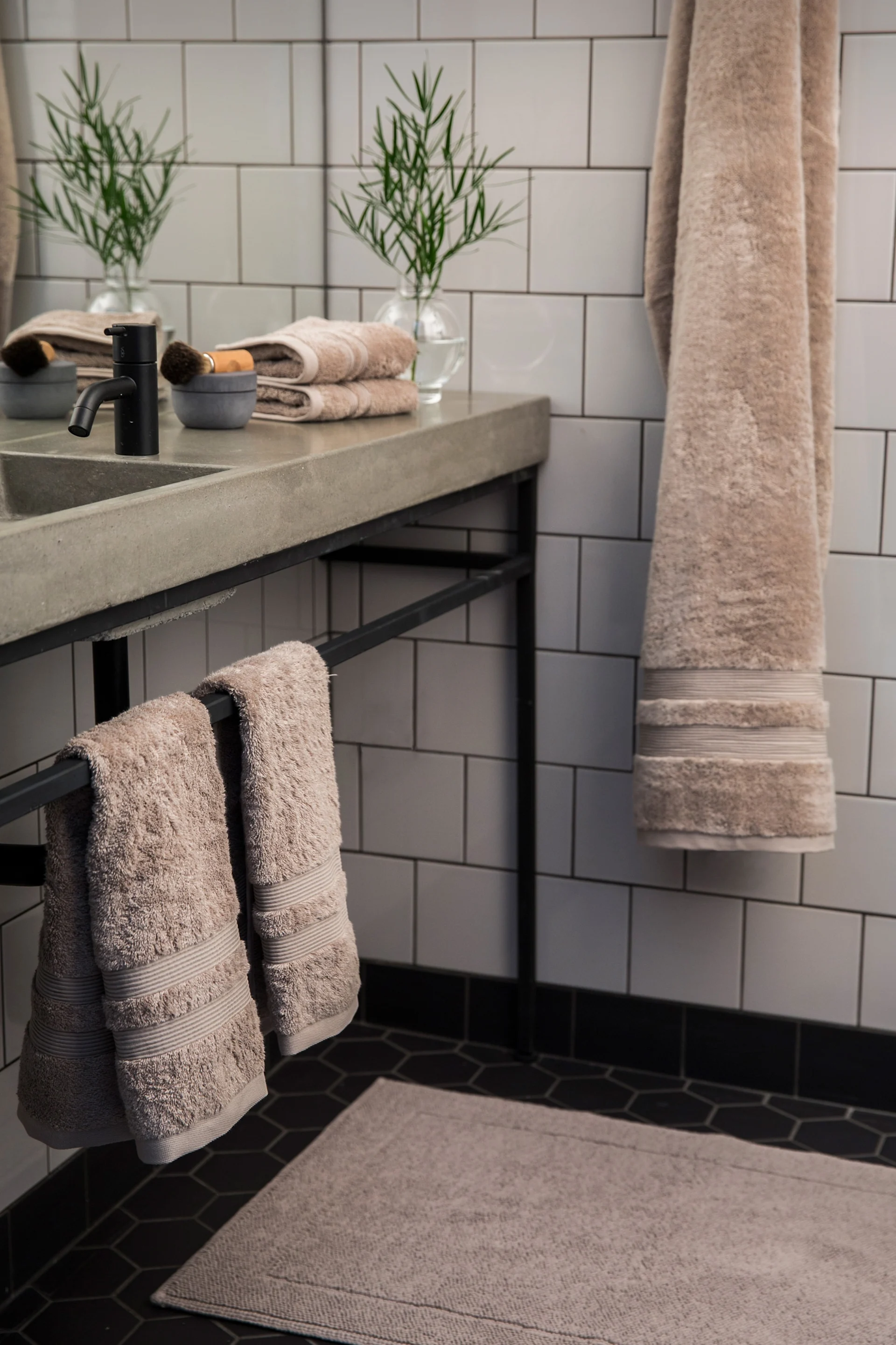 Modernes Bad mit Betonwaschbecken, schwarzem Wasserhahn, grüner Pflanze und beigen Handtüchern an Haltern und Haken. Schwarze Hexagonfliesen.